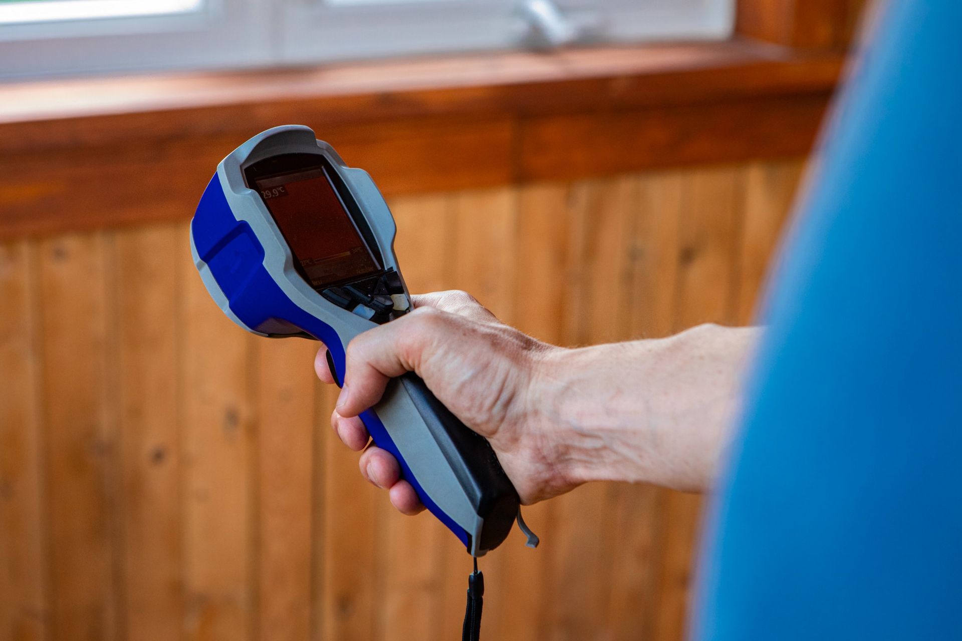 Person holding a blue and gray thermal camera pointed towards a wooden wall.