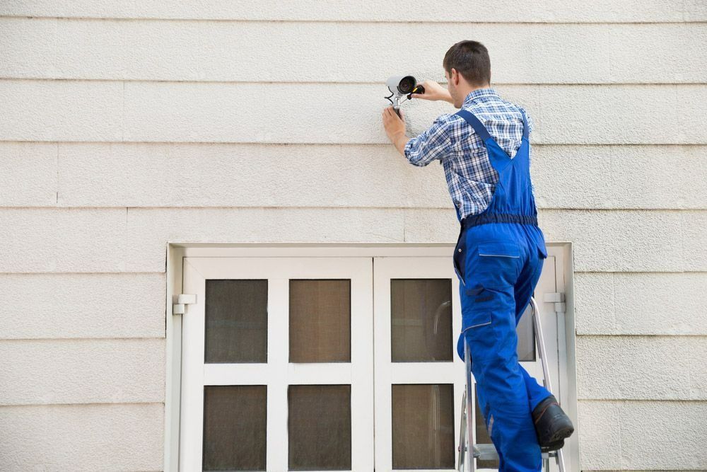 A Man Is Standing On A Ladder Installing A Camera On The Side Of A Building — On Guard Security & Communications In Wallsend, NSW