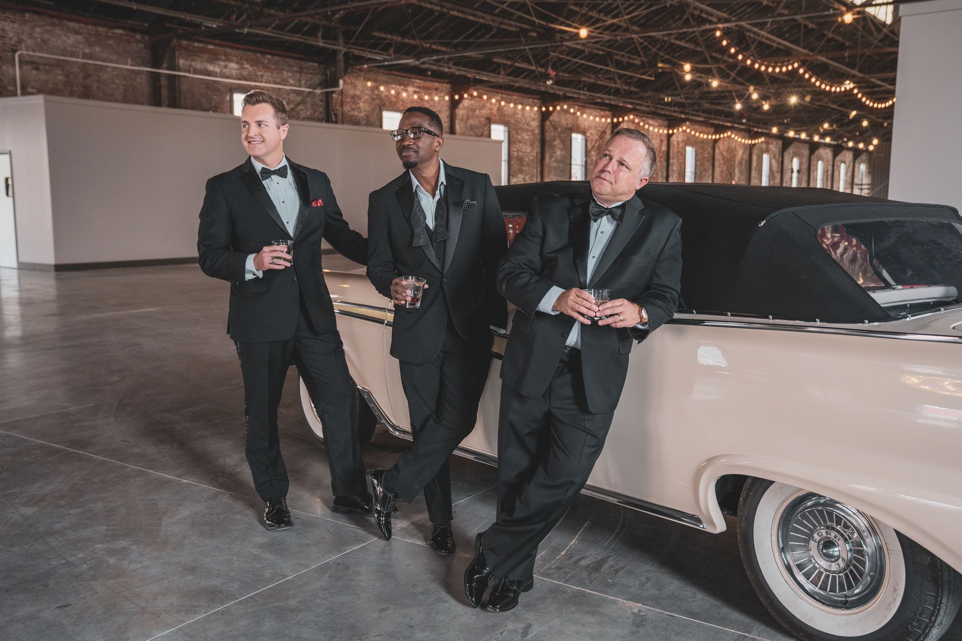 Three men in tuxedos are standing next to a car in a warehouse.