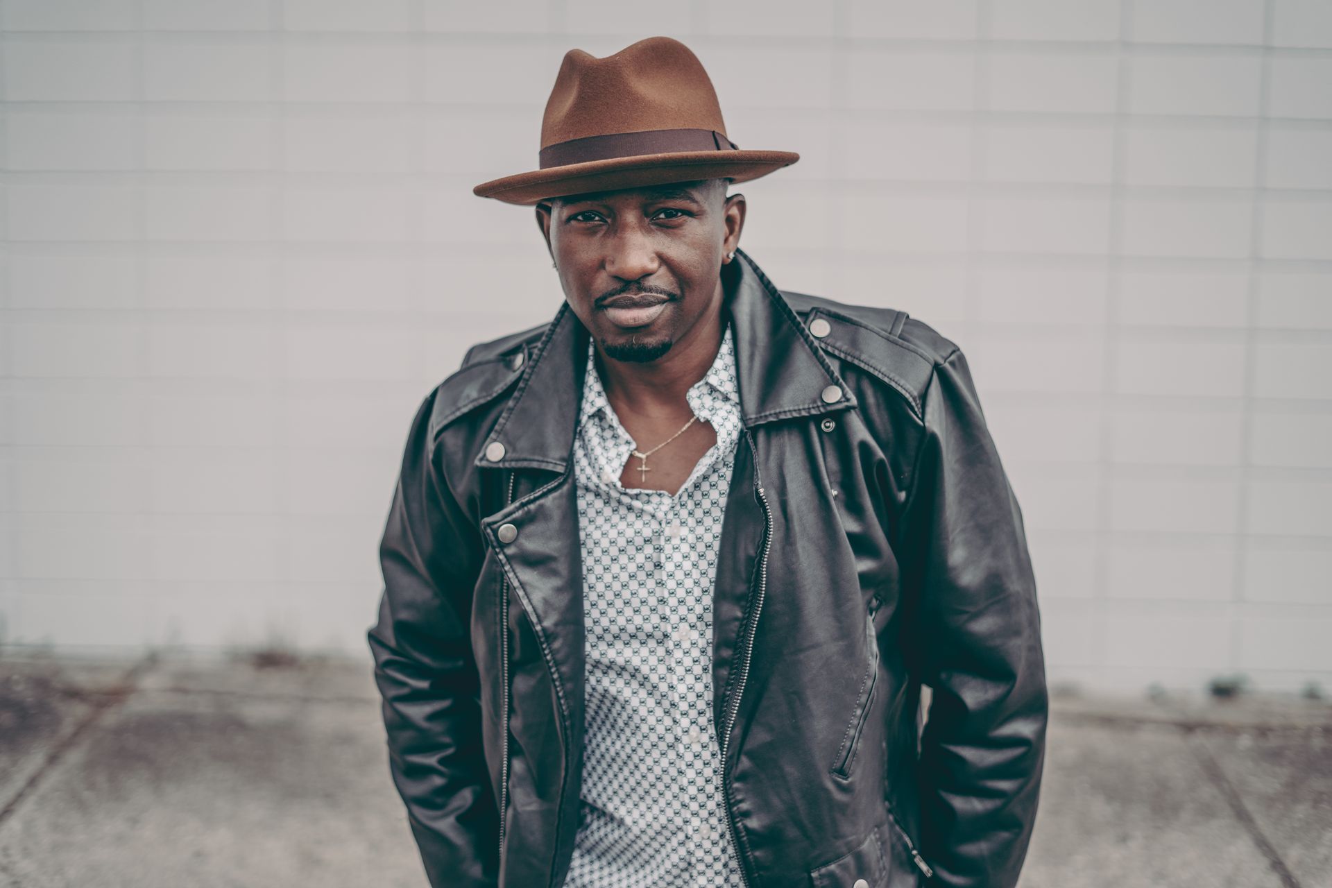 A man wearing a hat and a leather jacket is standing in front of a white wall.