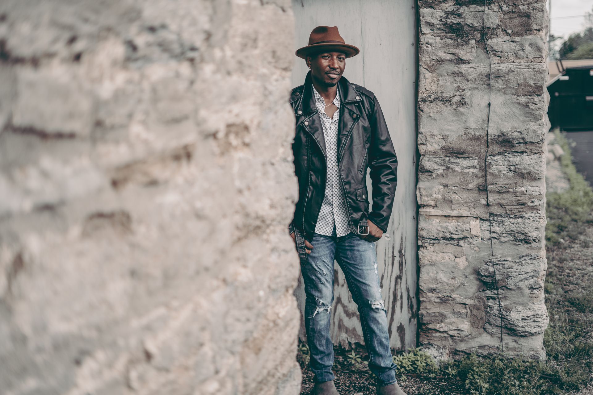 A man wearing a hat and a leather jacket is leaning against a brick wall.