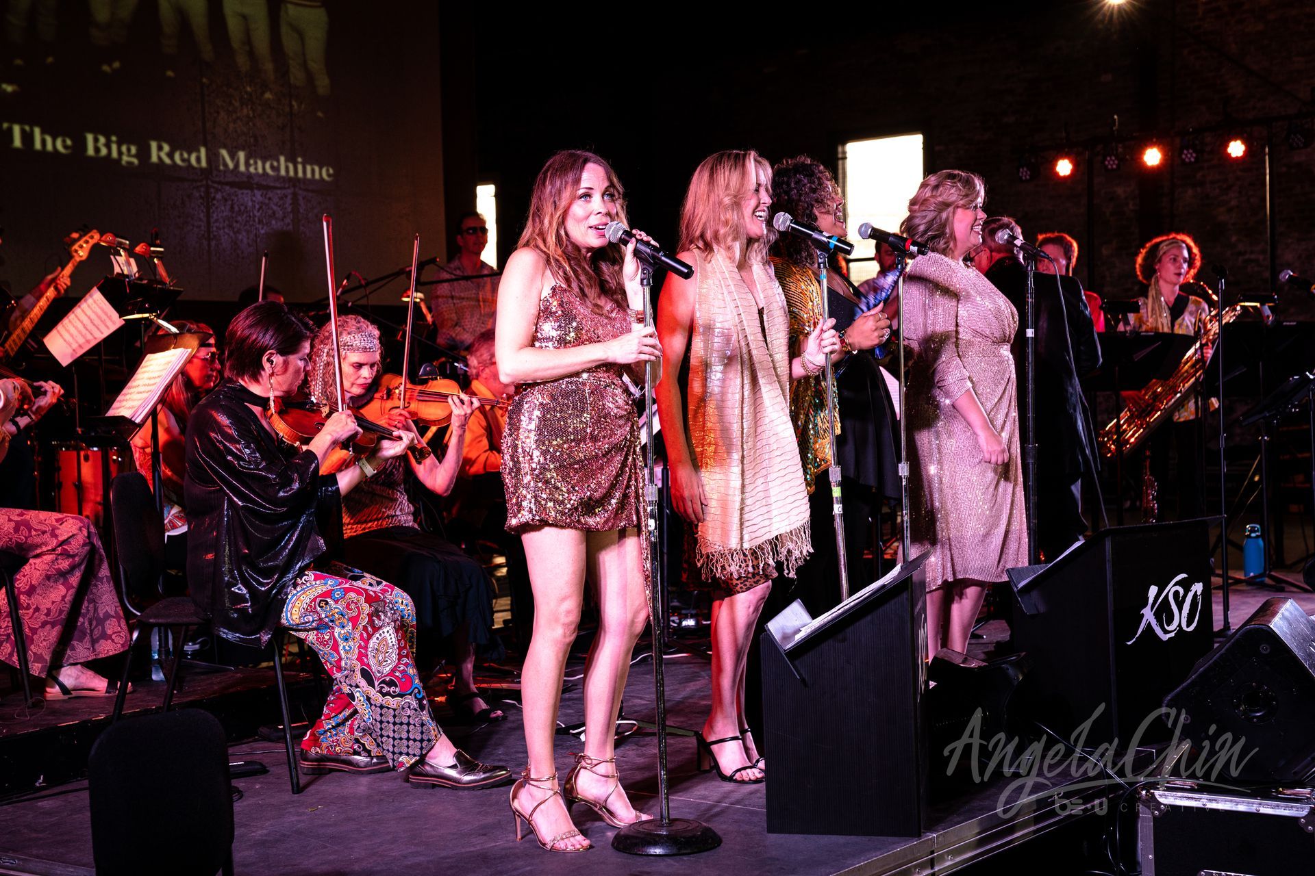 A group of women are singing on a stage in front of an orchestra.