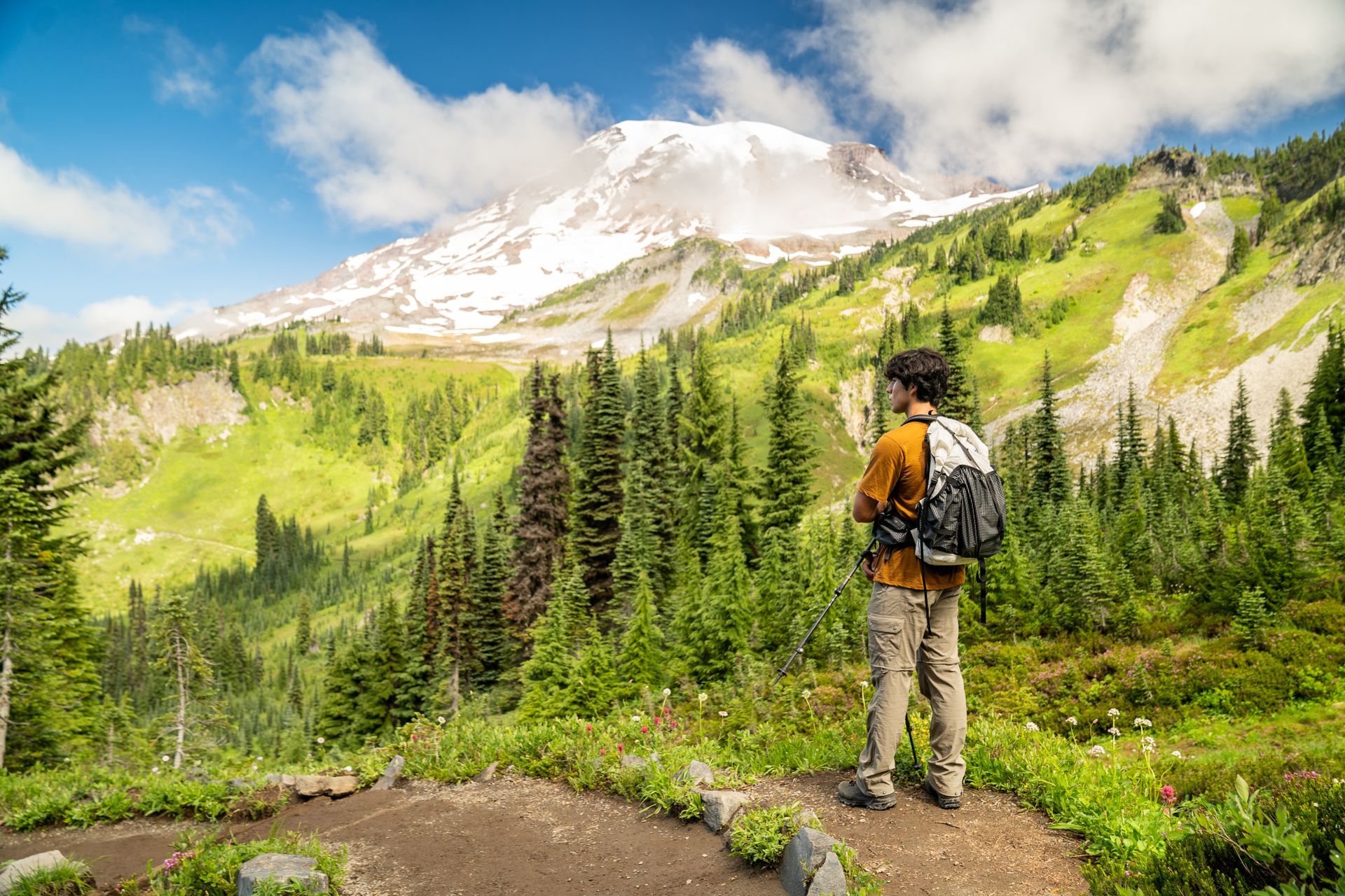 A man with a backpack is standing on a trail looking at a mountain.