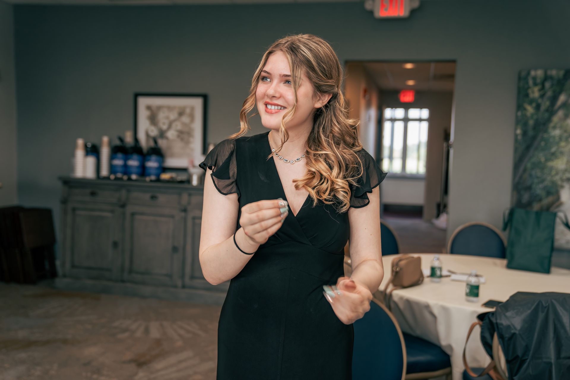 A woman in a black dress is standing in a room with a table and chairs.