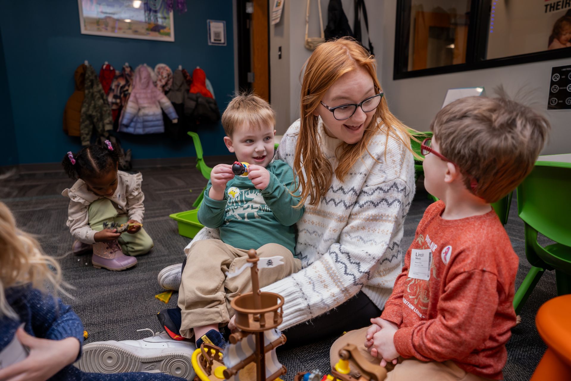 A woman is sitting on the floor with a group of children playing with toys.