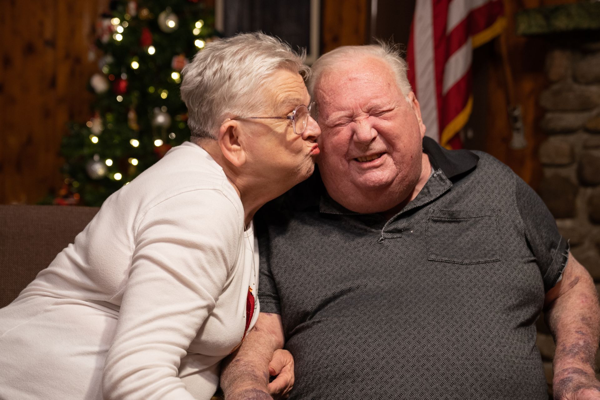 A woman is kissing a man on the cheek in front of a christmas tree.