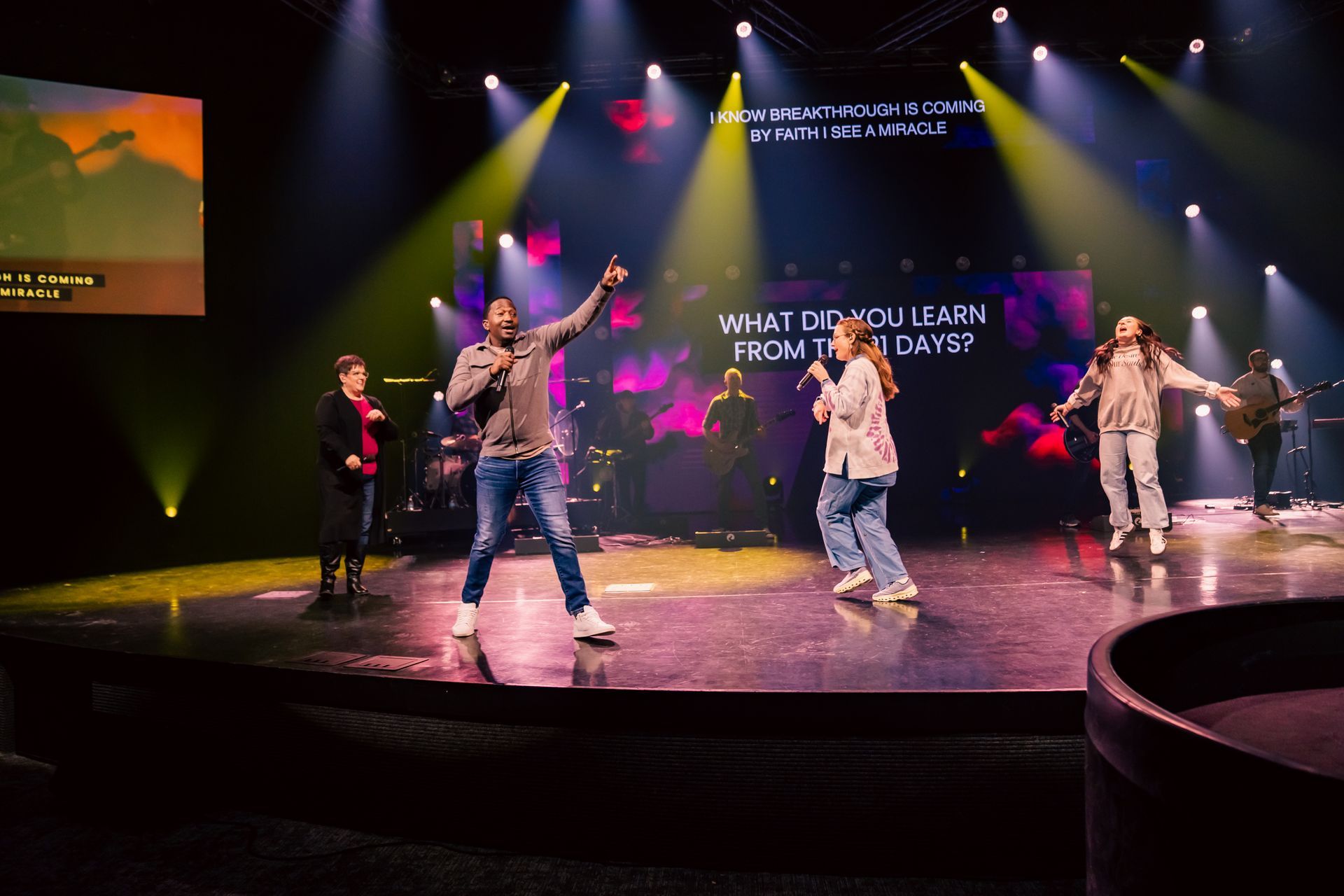 A group of people are dancing on a stage in front of a large screen.