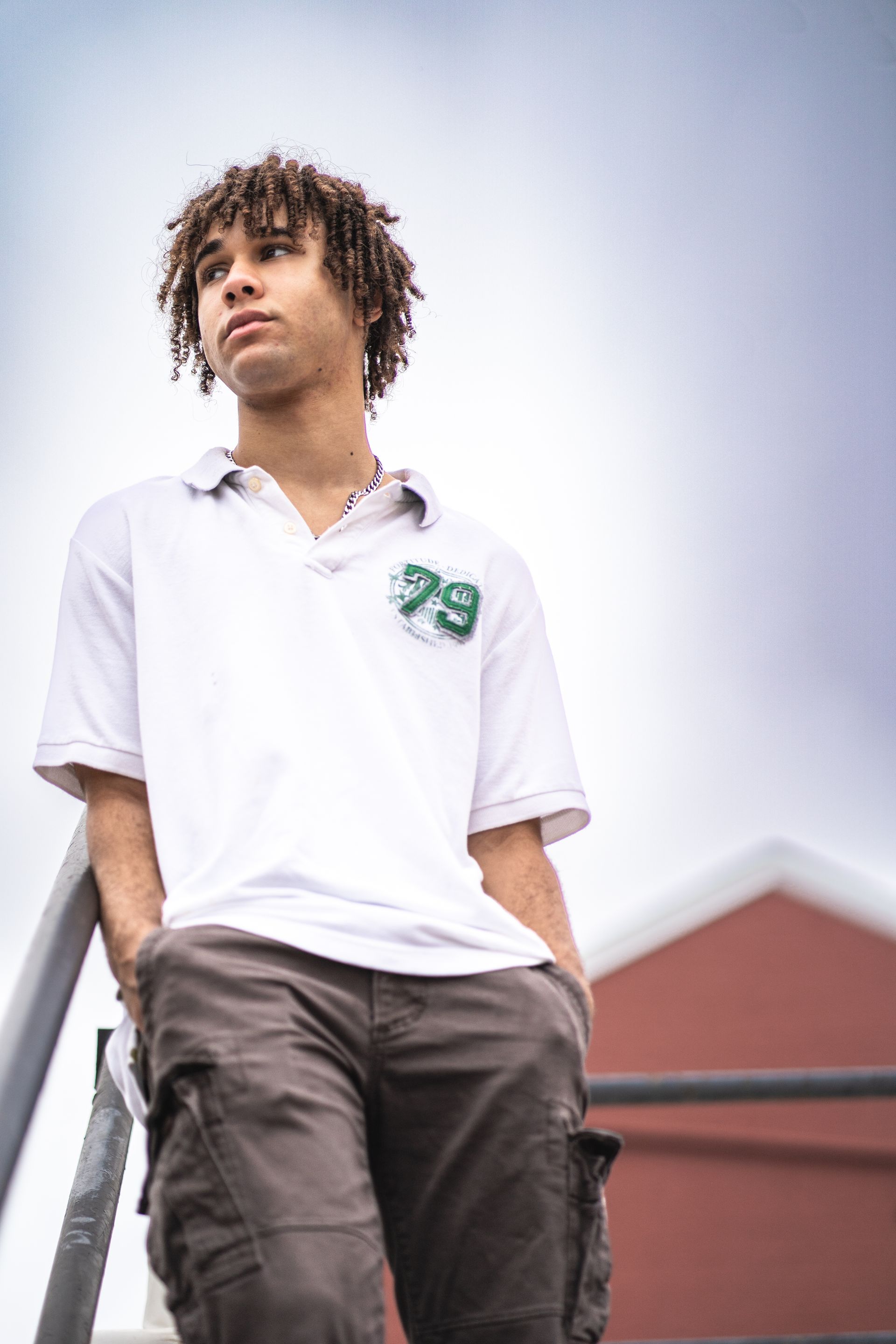 A young man in a white shirt and brown pants is leaning on a railing.