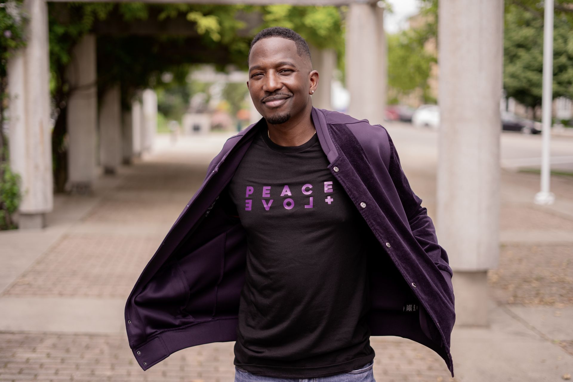 A man wearing a black shirt and a purple jacket is standing on a sidewalk.