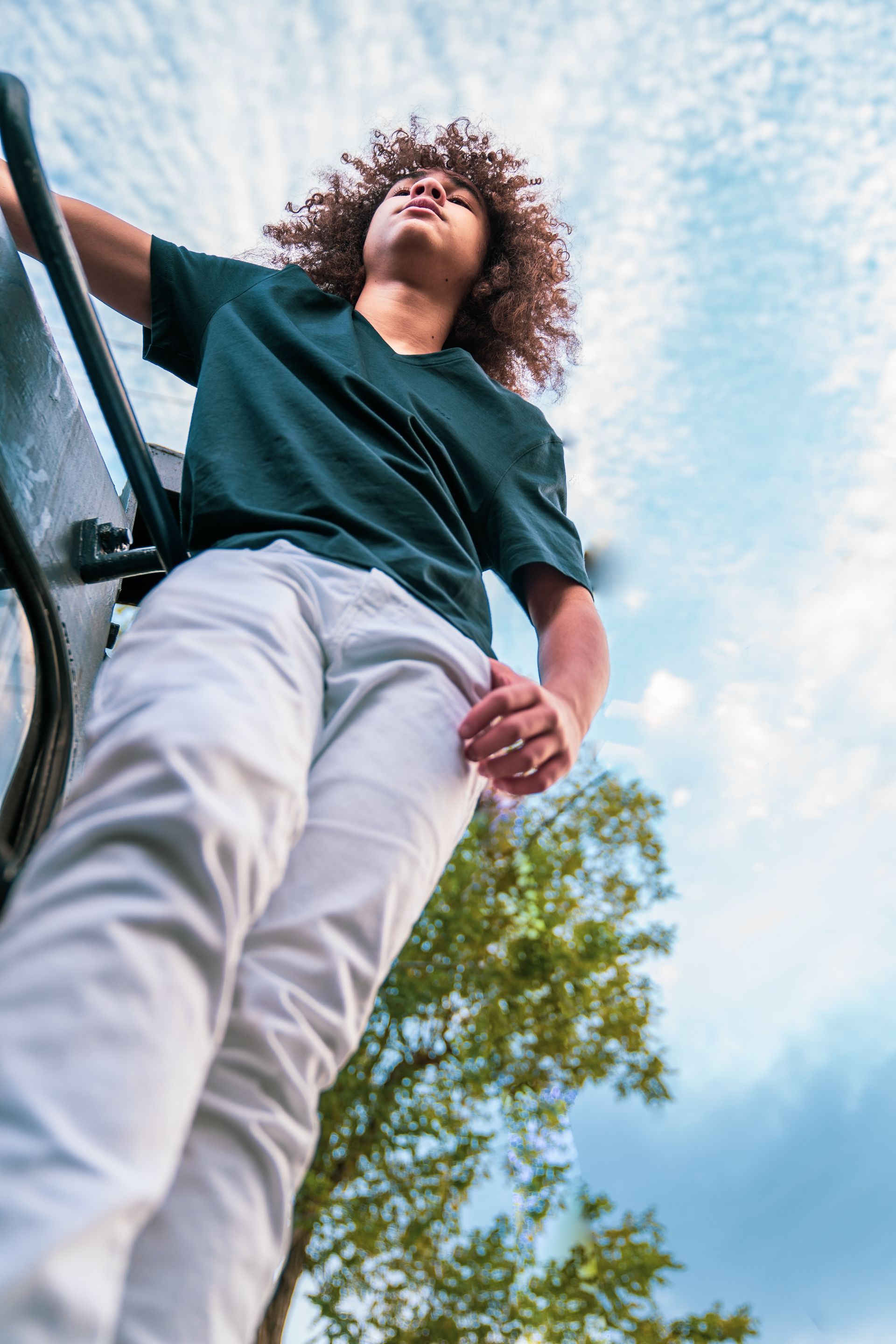 A young man with curly hair is standing on a ladder looking up at the sky.