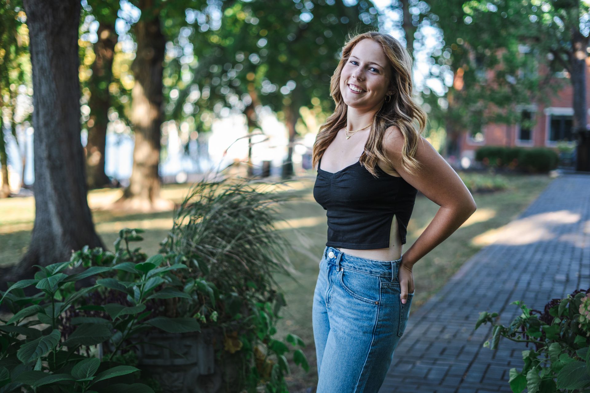 A woman in a black tank top and blue jeans is standing on a sidewalk in a park.