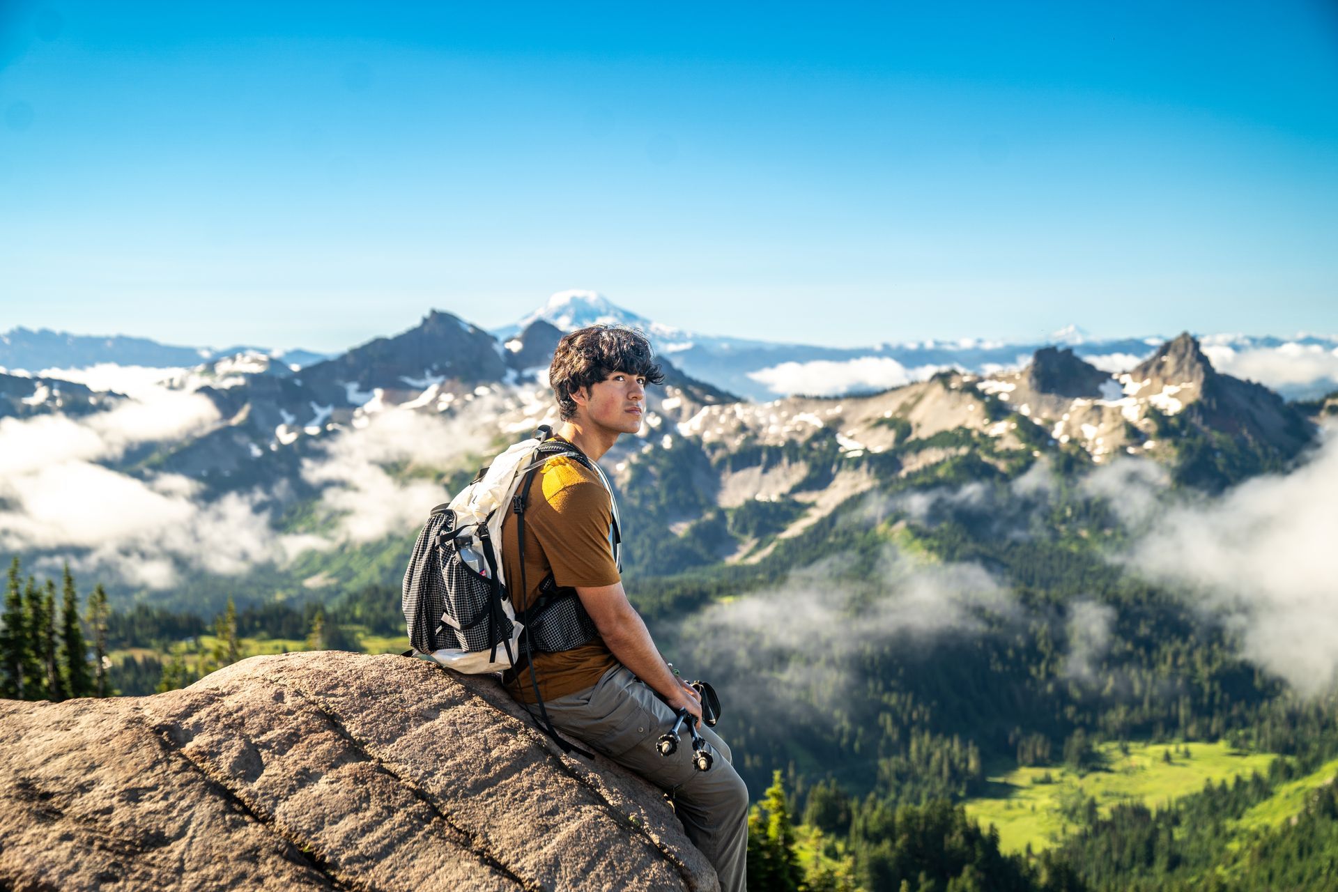 A man with a backpack is sitting on top of a rock overlooking a mountain range.