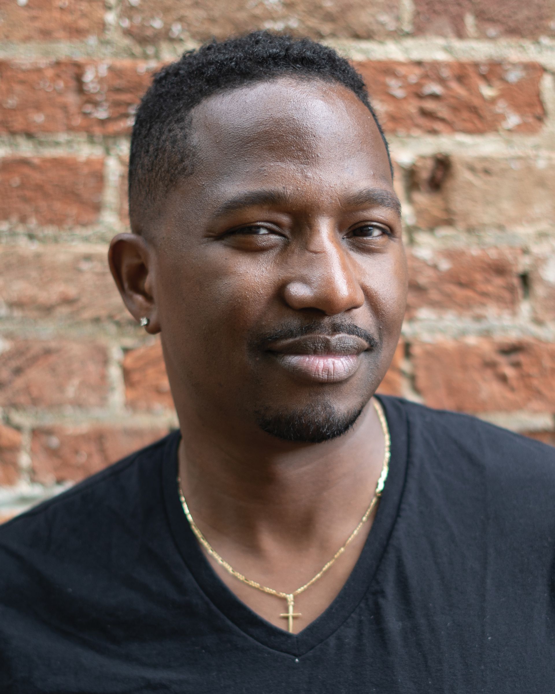 A man wearing a black shirt and a gold cross necklace is standing in front of a brick wall.