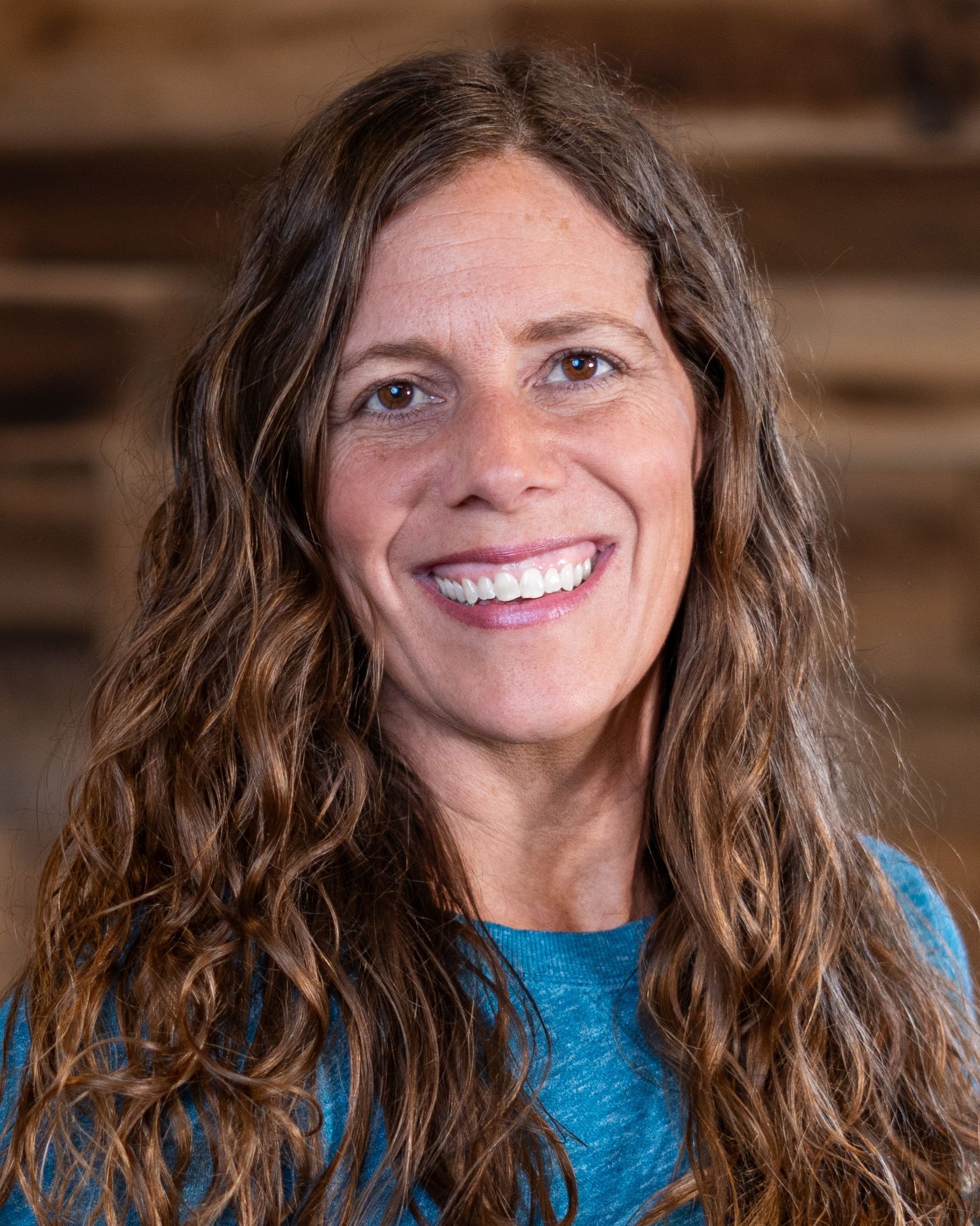 A woman with long curly hair is smiling for the camera while wearing a blue shirt.