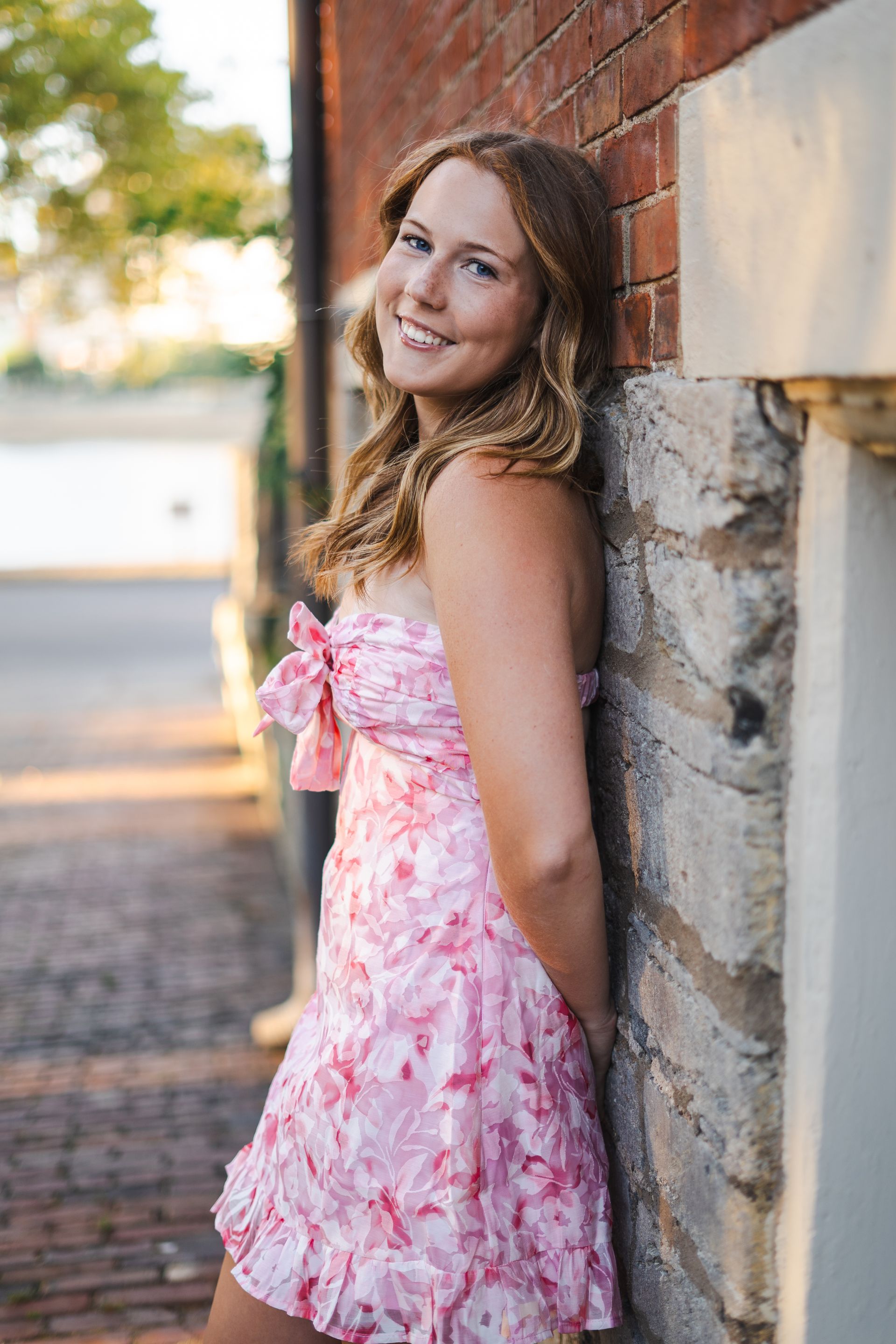 A woman in a pink dress is leaning against a brick wall.
