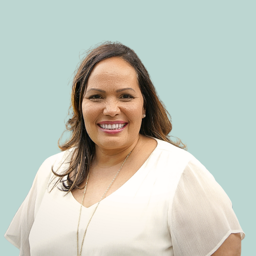 Woman smiling, resting chin on hand. Background shows a home office with desk and bookshelves.