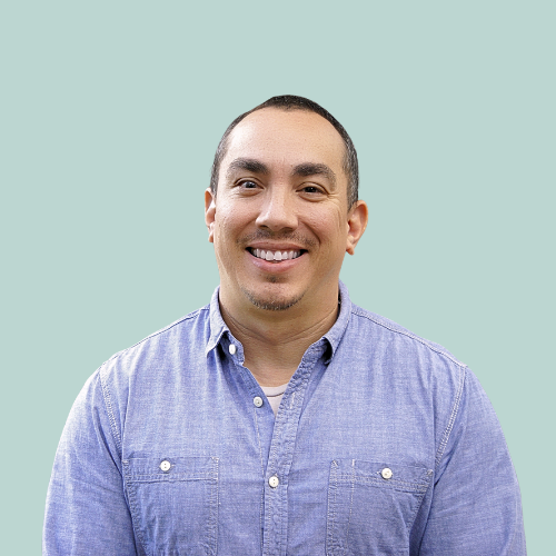 Smiling man wearing a blue button-down shirt. DJ equipment and headphones are in the background.