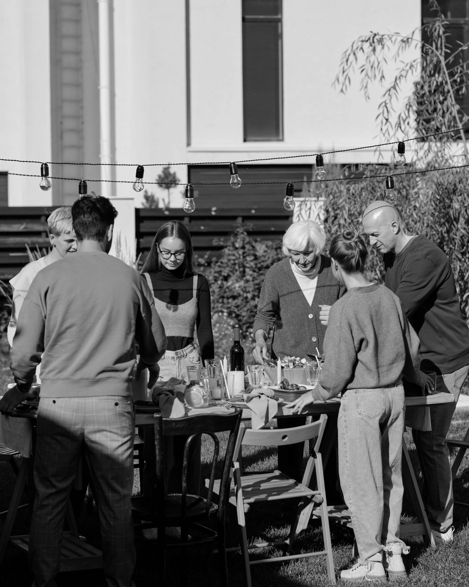 People gathered around a table outdoors beside a building, chatting and eating.