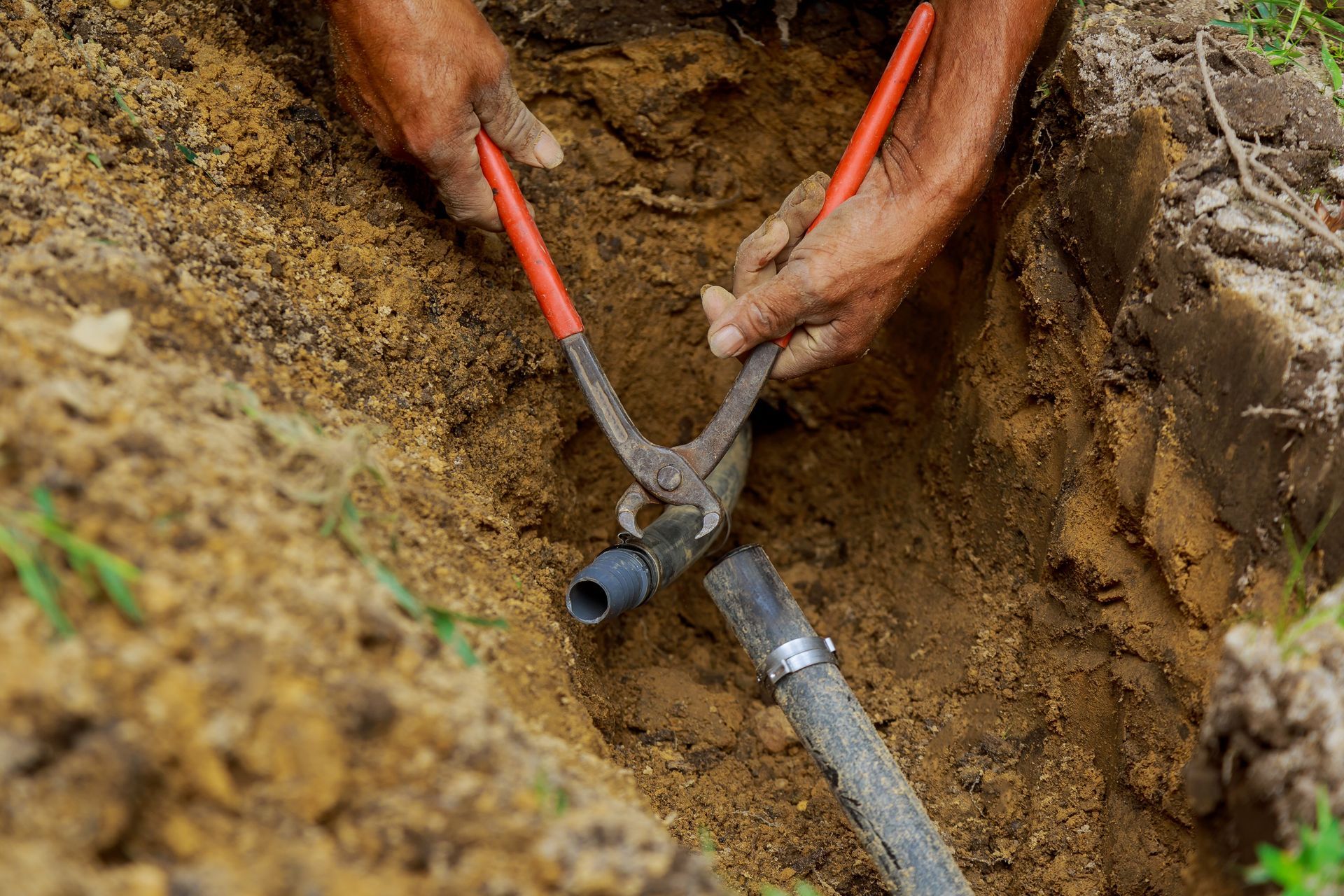 Hands using orange-handled pliers to work on a pipe buried in a dirt trench.