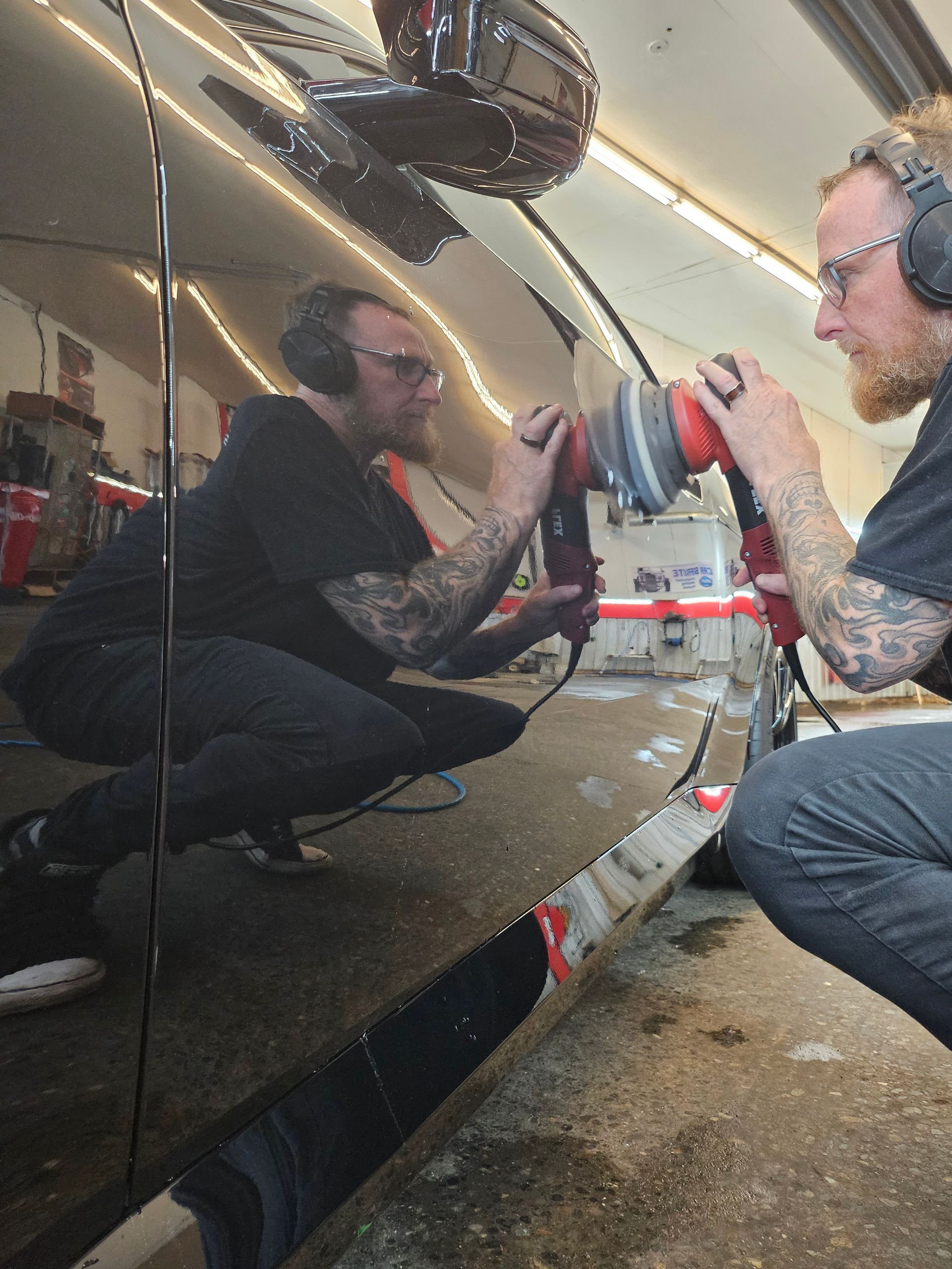 Man with tattoos polishes a black car, wearing earmuffs. Garage setting.