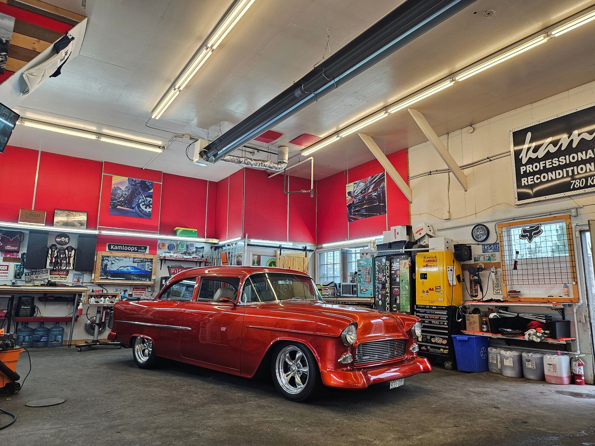 Shiny orange classic car inside a well-lit garage with tools, shelves, and red walls.