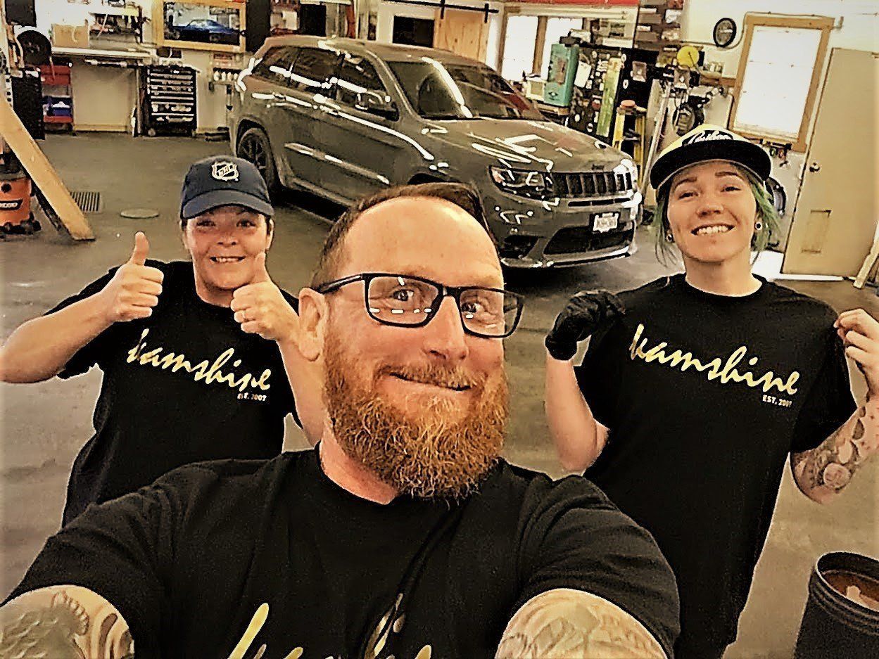 Three people in a garage pose with a grey SUV. They wear black shirts, one gives a thumbs up.