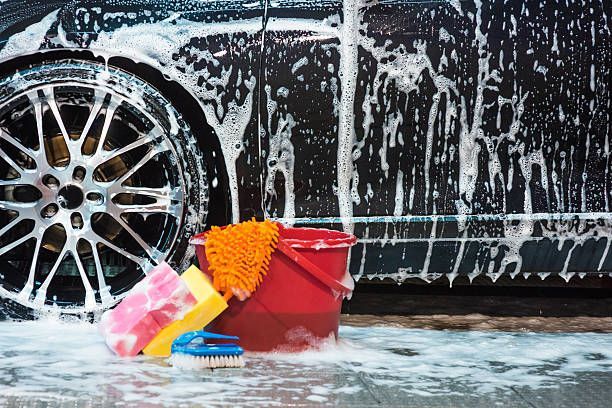 Black car being washed with soapy water, bucket, sponges, and brush.