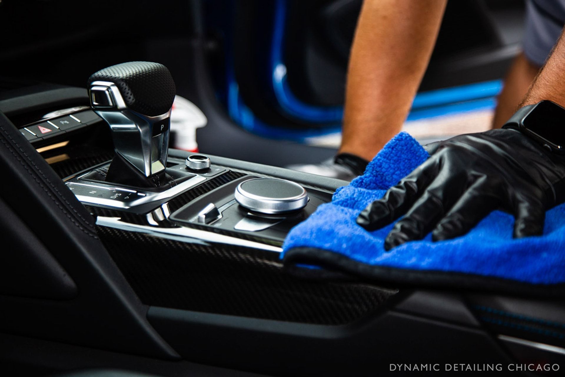 Gloved hand wiping a car's center console with a blue microfiber cloth; detailing interior.