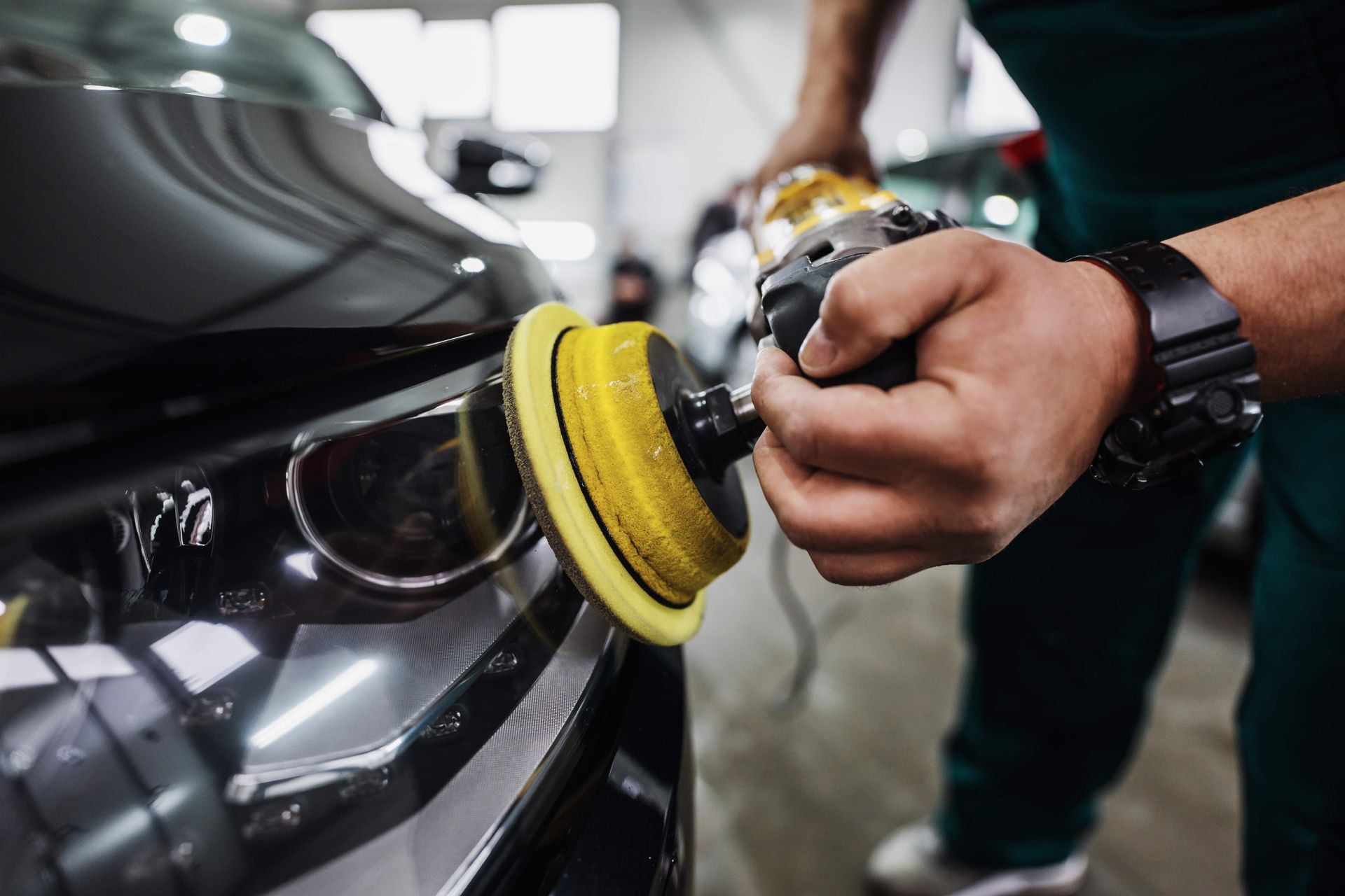 Person polishing a black car headlight with a yellow buffing pad in a garage.