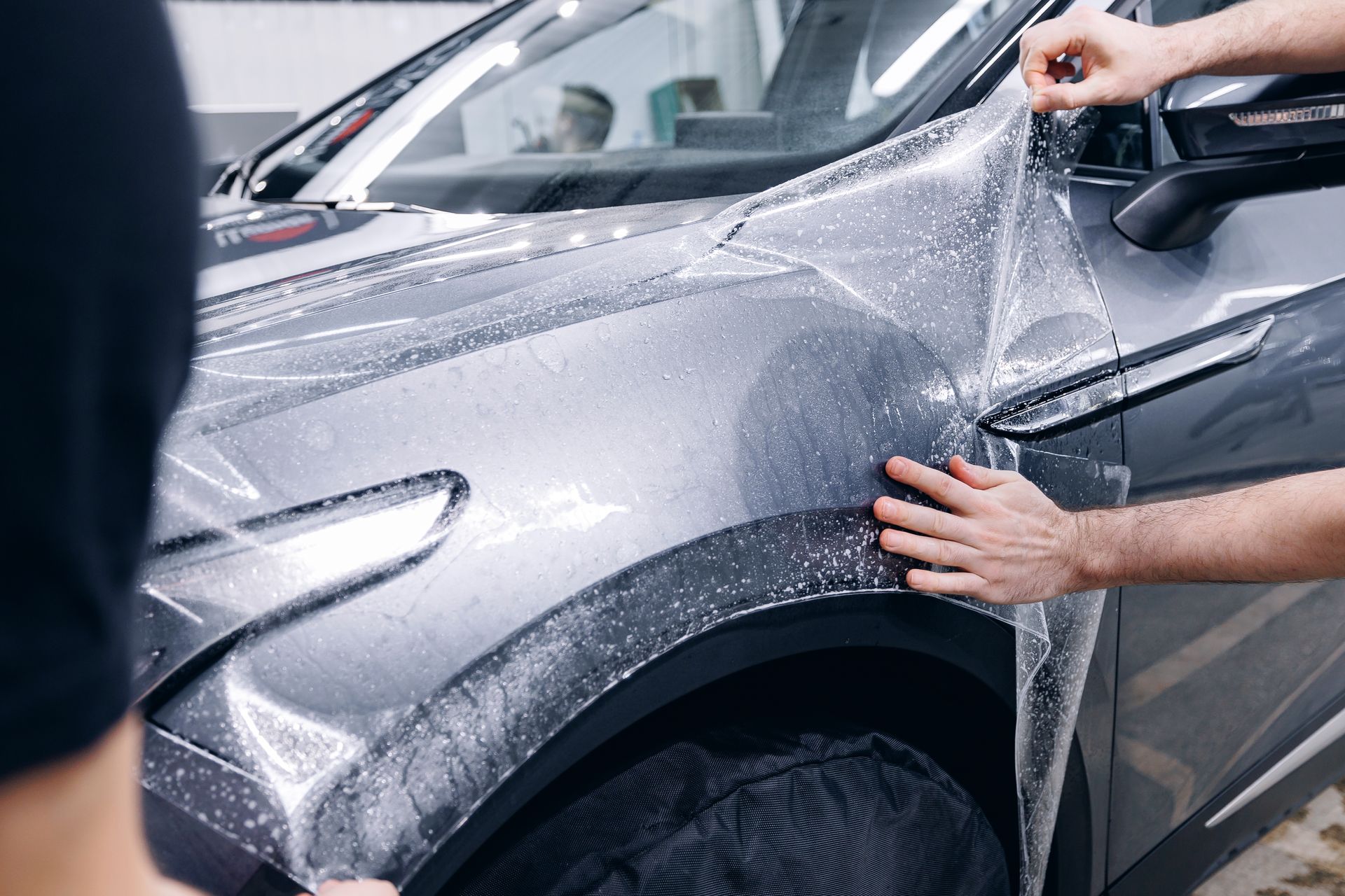 Applying protective film to a gray car's fender. Two hands carefully smoothing out the clear film.