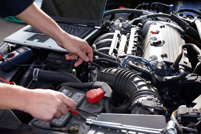 A person is working on a car engine with a laptop in the background