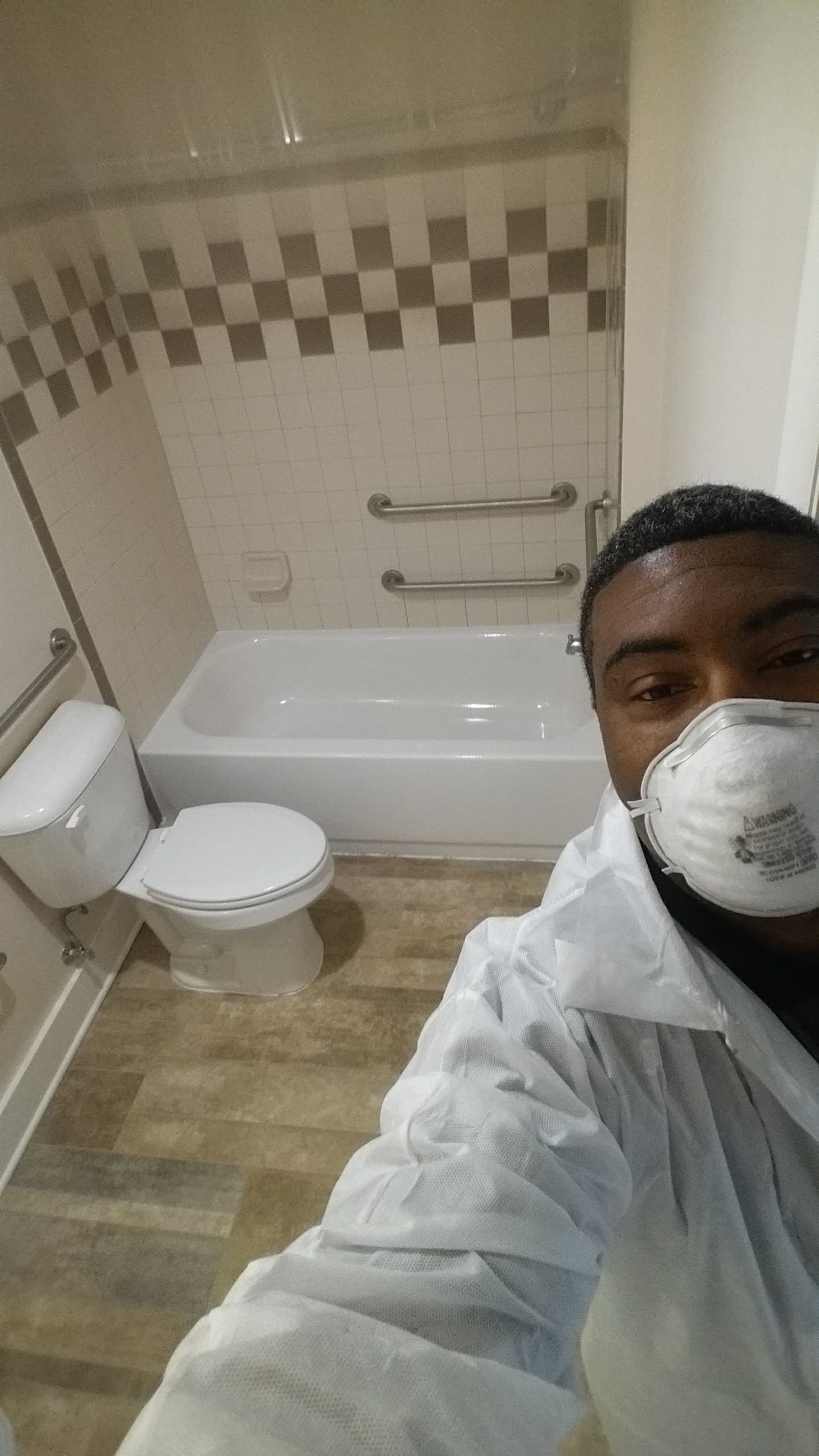 Person in a protective suit and mask in a bathroom with a tub, toilet, and tile wall.