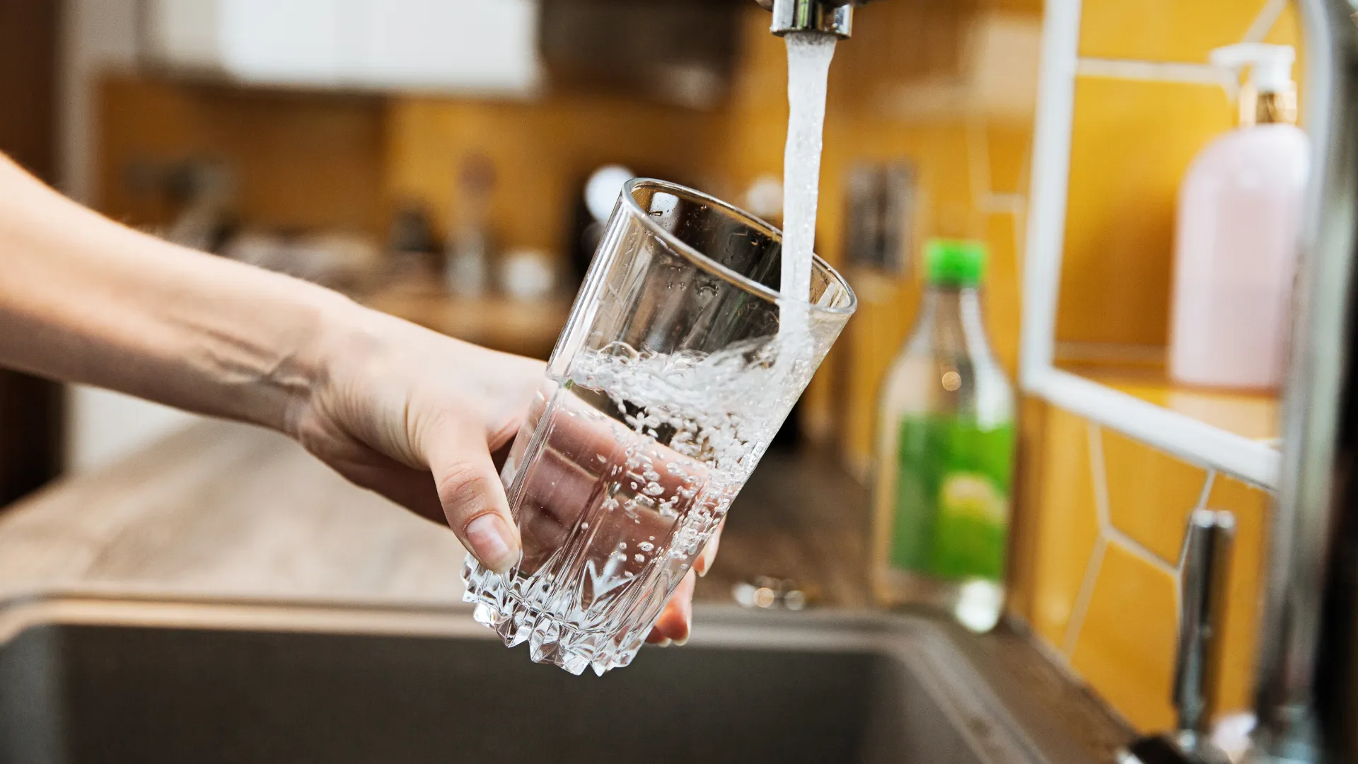 A hand fills a clear, textured glass with water from a kitchen faucet against a background of yellow tiles.
