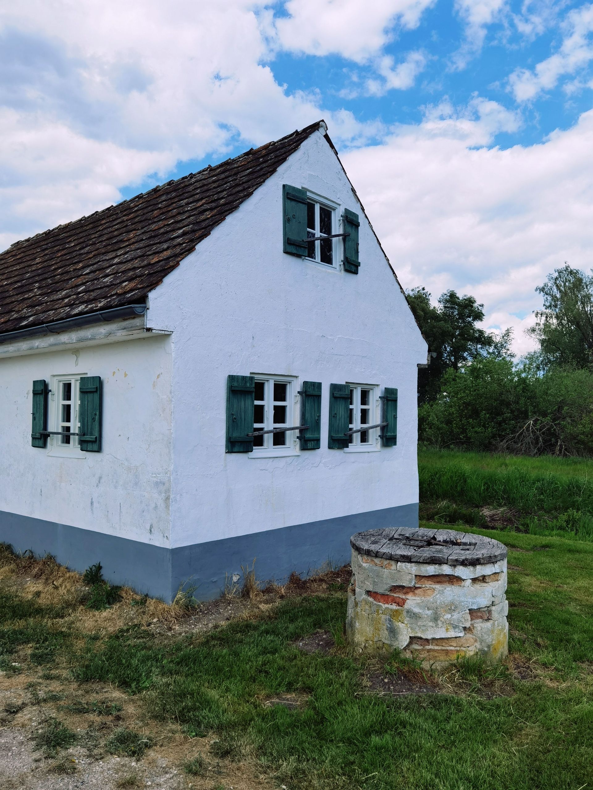 White cottage with green shutters and a steep roof beside an old stone well under a cloudy sky