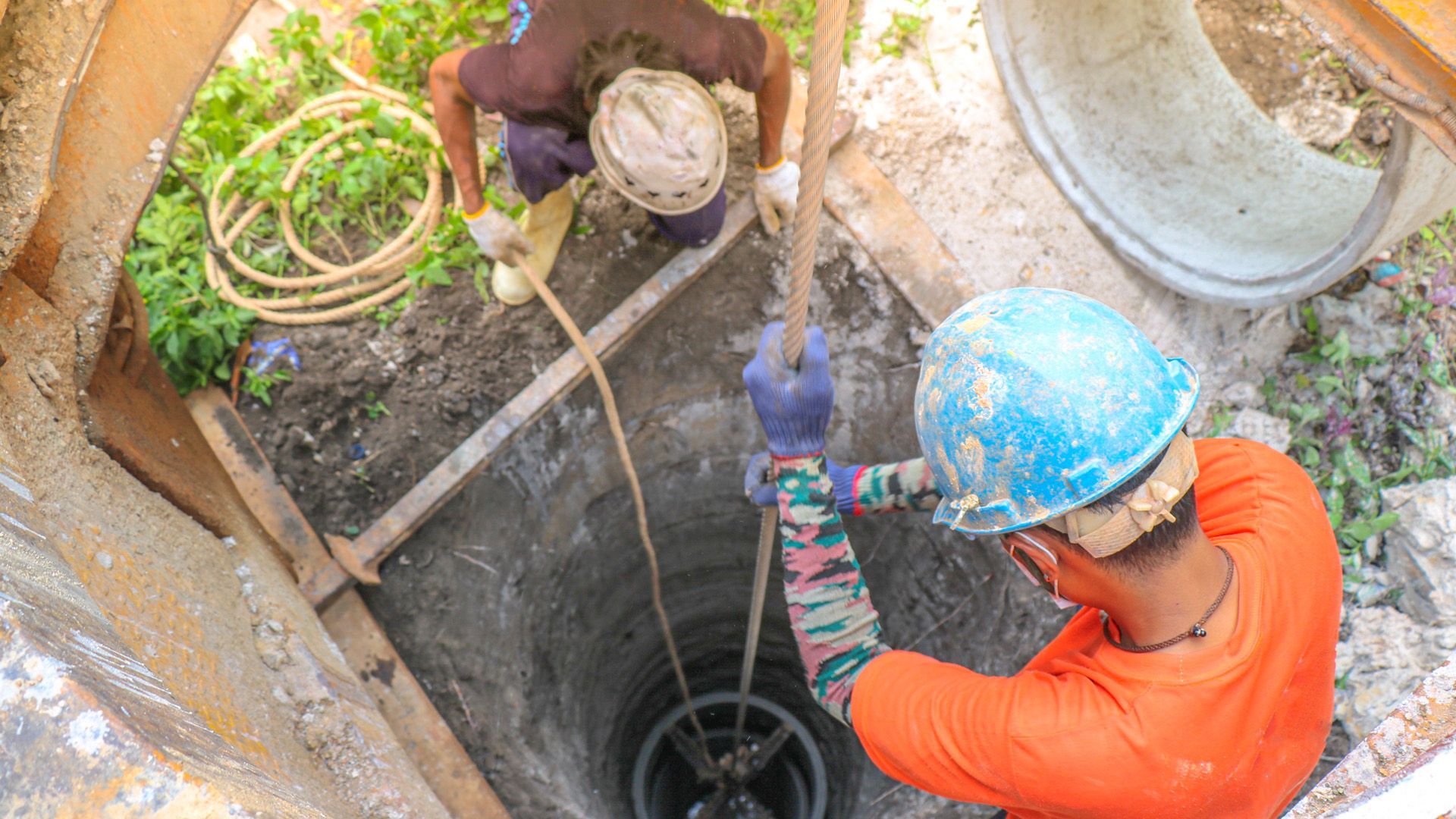Two workers inside and above a well, one pulling a rope, one securing it, orange and blue shirts.