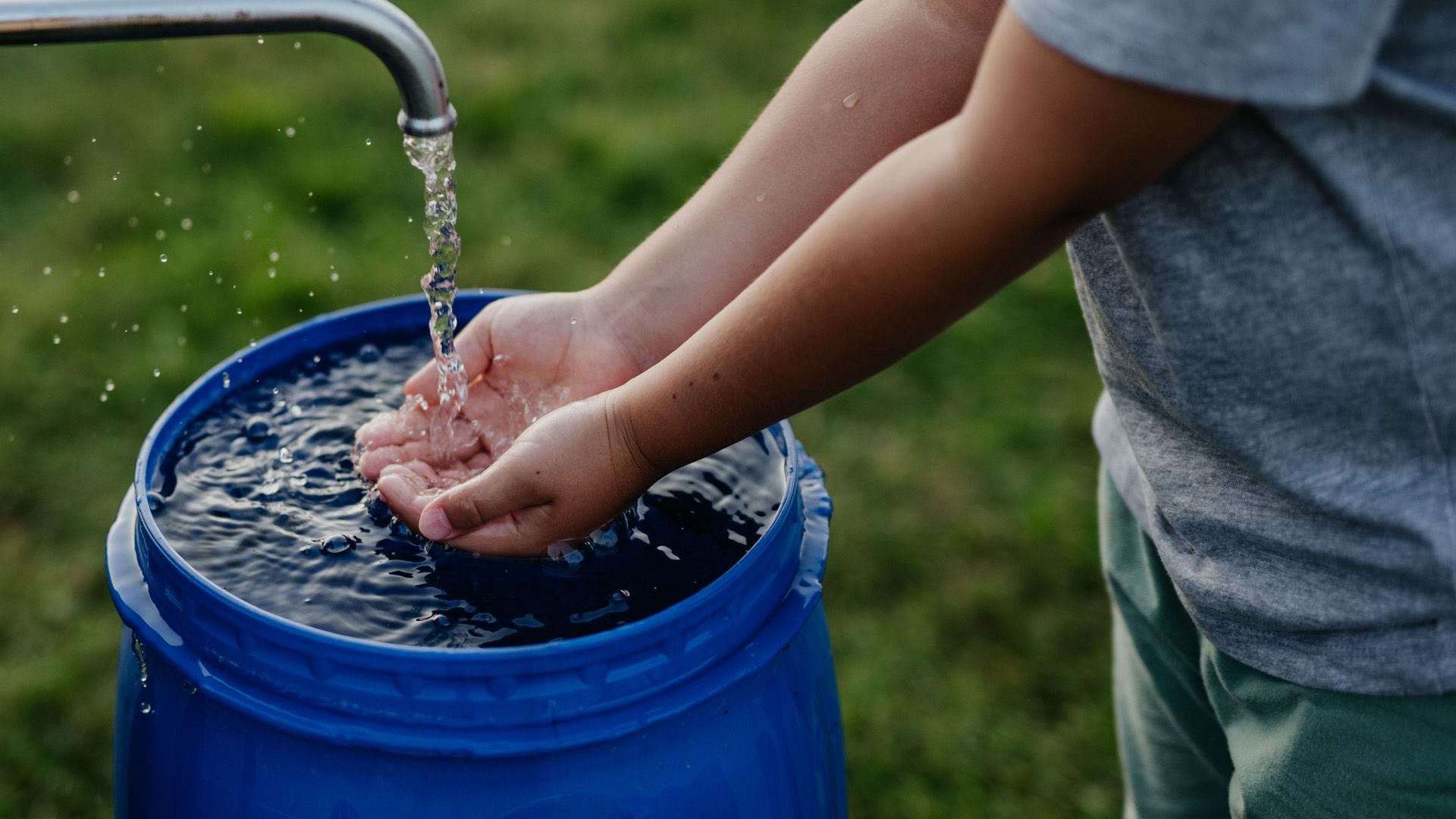 Child washing hands with water flowing into a blue barrel outdoors.