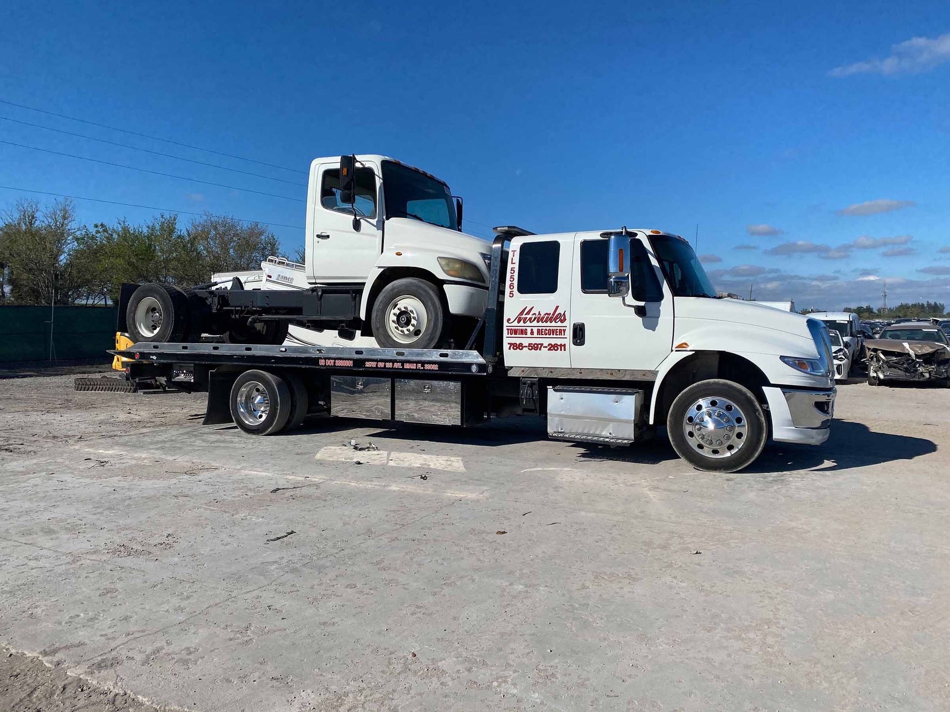 White tow truck carrying a cab-over truck on its flatbed, under a bright, sunny sky.