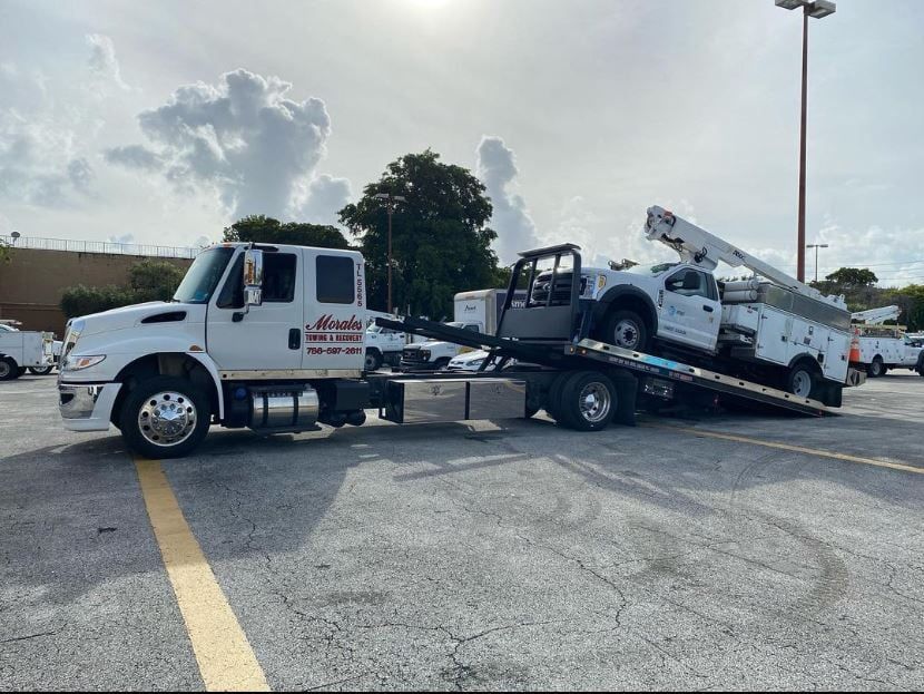 A white tow truck is transporting a white utility truck with an extended boom on a sunny day.