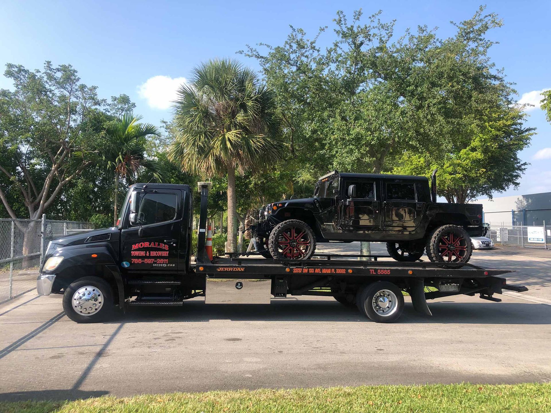 Black tow truck carrying a classic car with red rims, parked on pavement near trees.