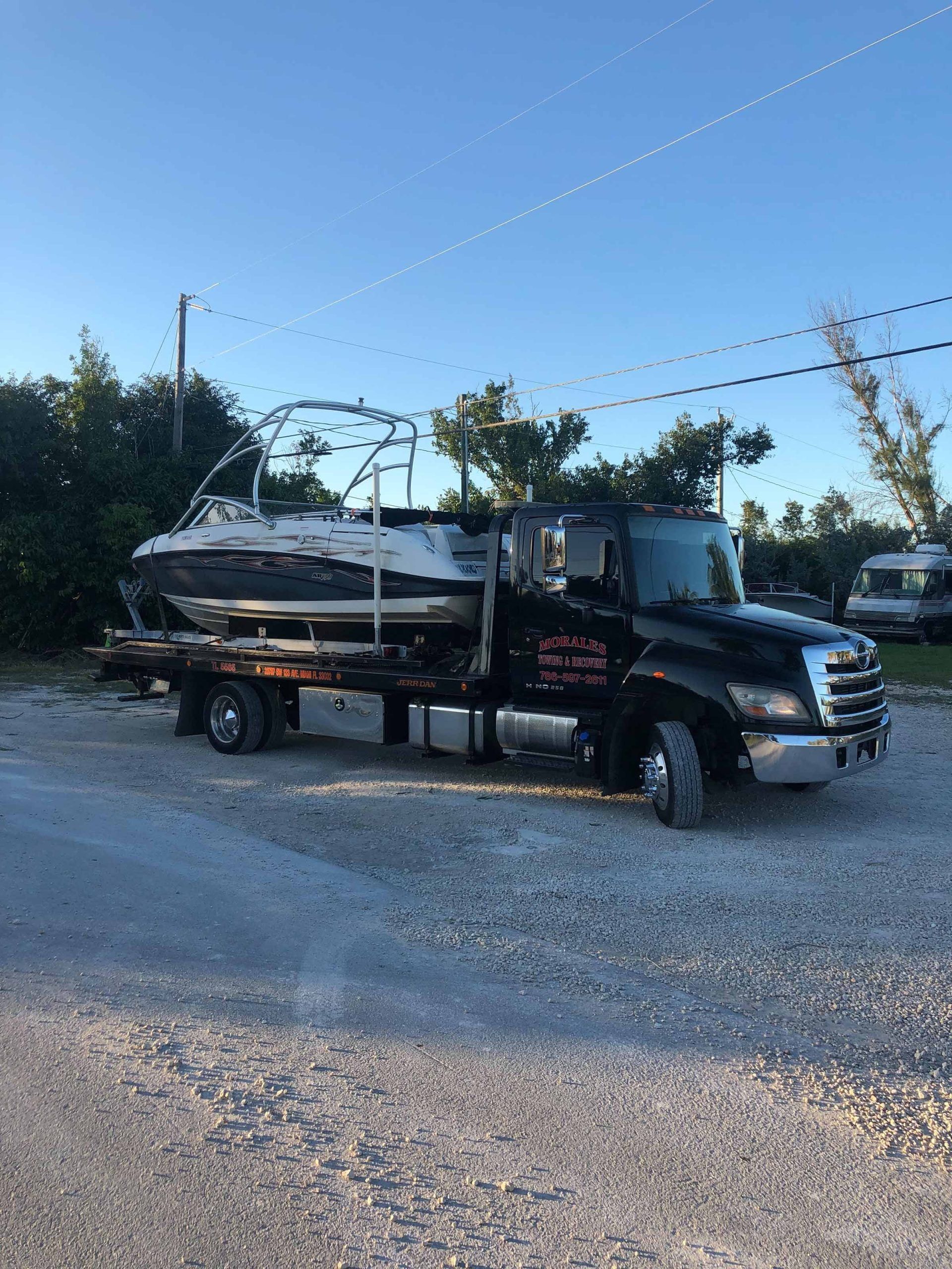 Black tow truck carrying a boat on a gravel road, trees in the background, blue sky.