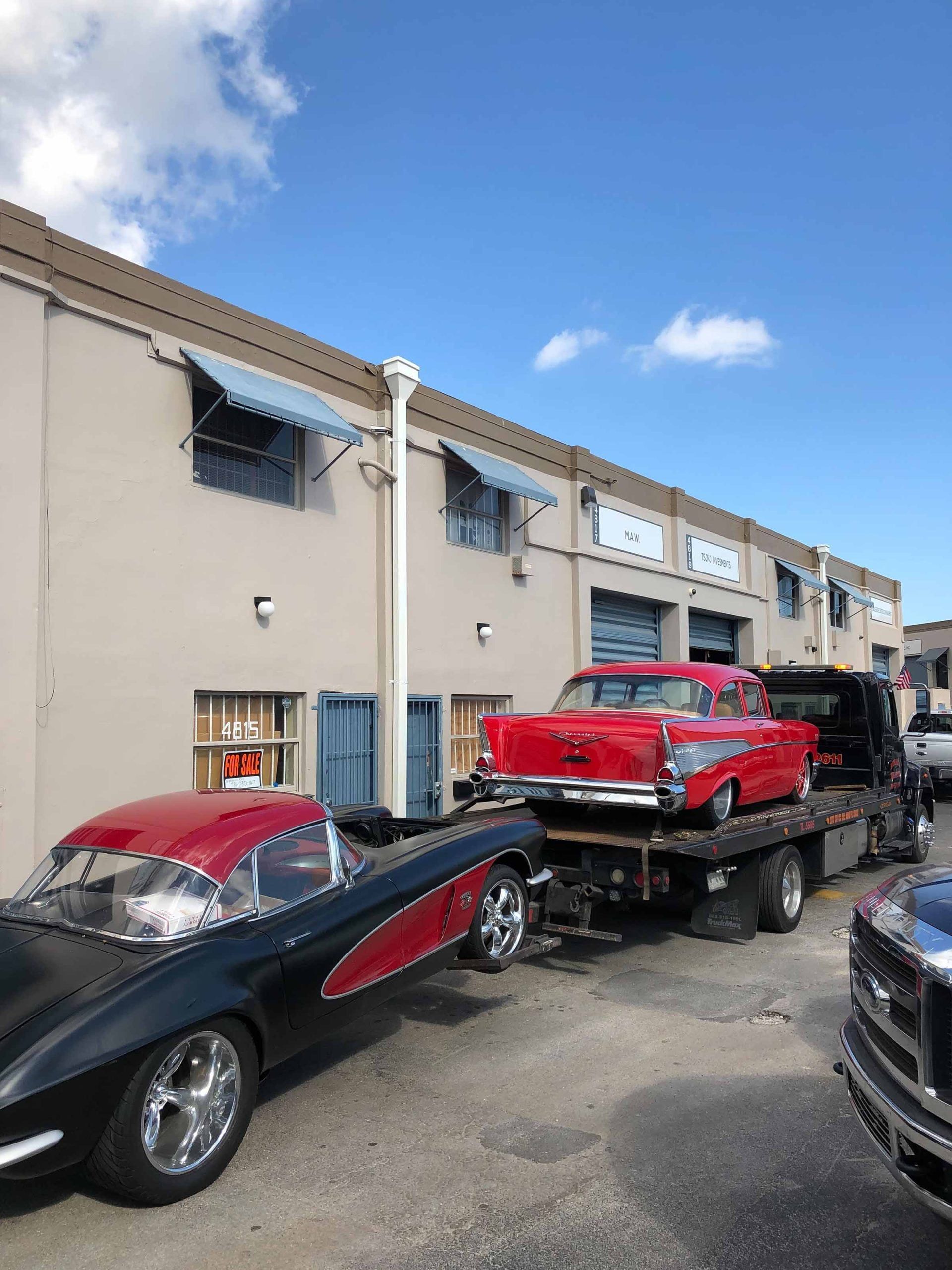 A black and red classic car beside a flatbed truck carrying a red classic car, parked near a building.