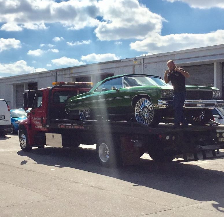 Green classic car on a tow truck, man standing beside it, sunny day.