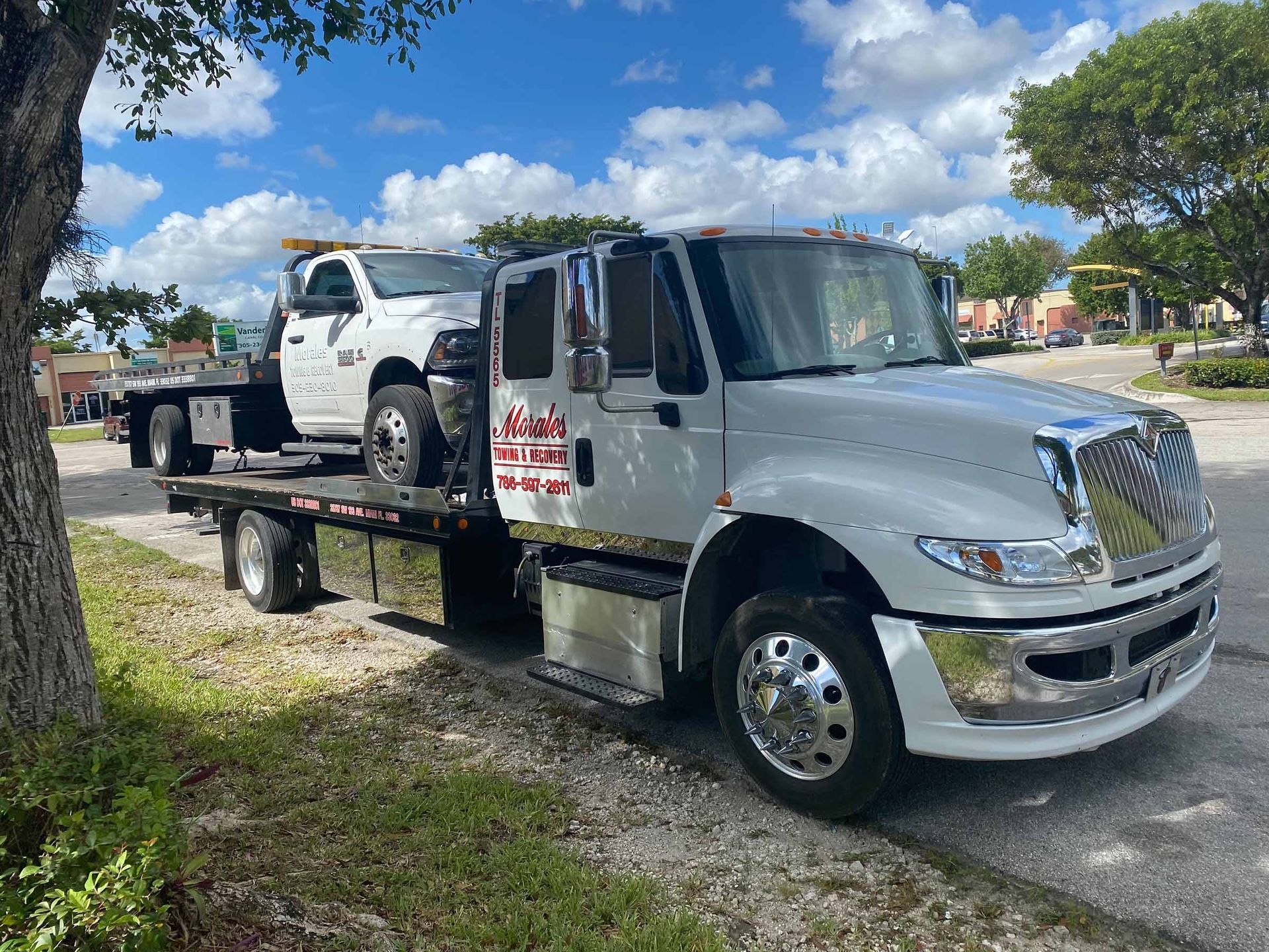 White tow truck with a smaller white truck on its flatbed, parked on a street near a tree.