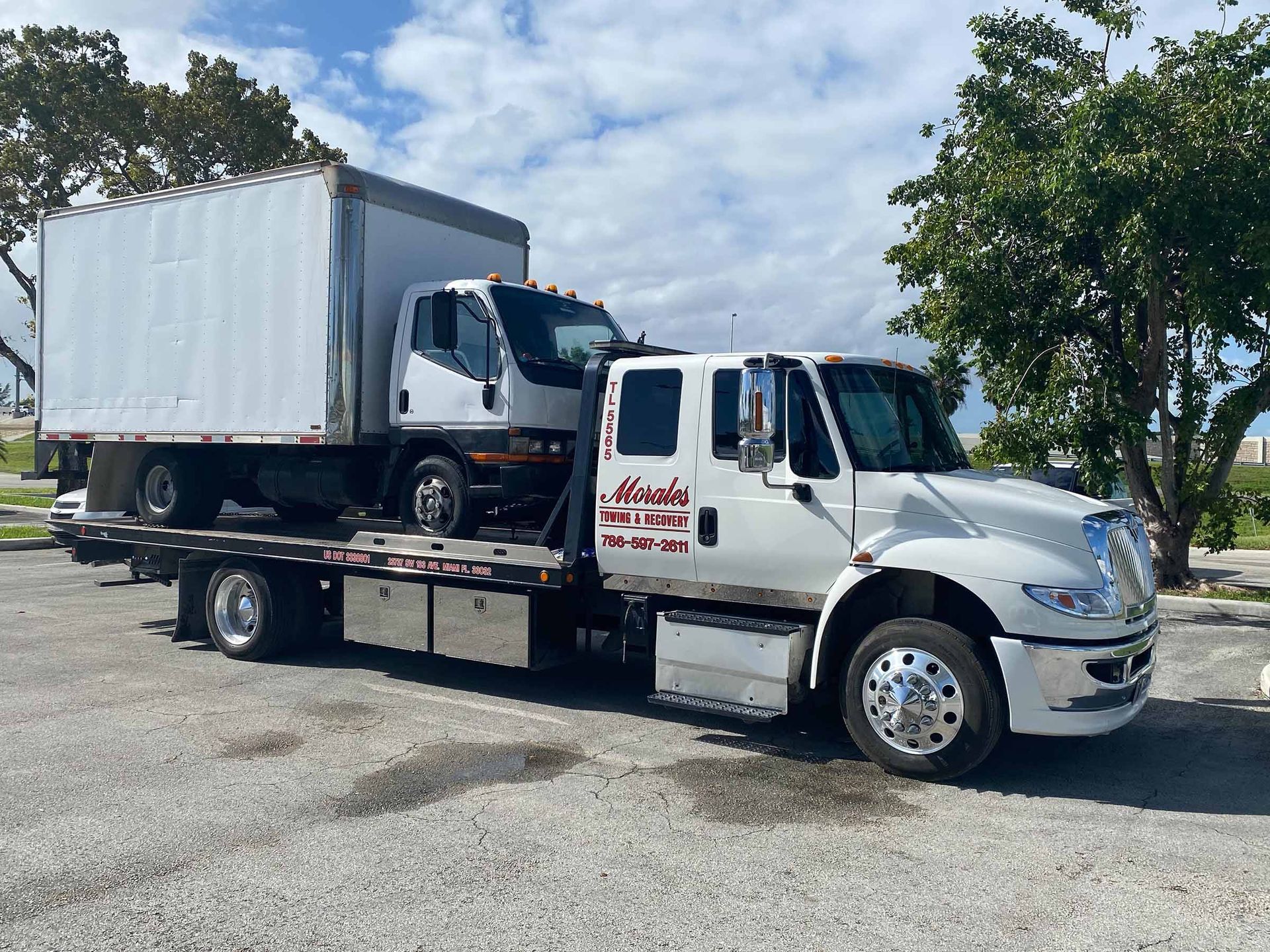 Tow truck carrying a box truck on a flatbed, parked outdoors. Cloudy sky, asphalt surface.