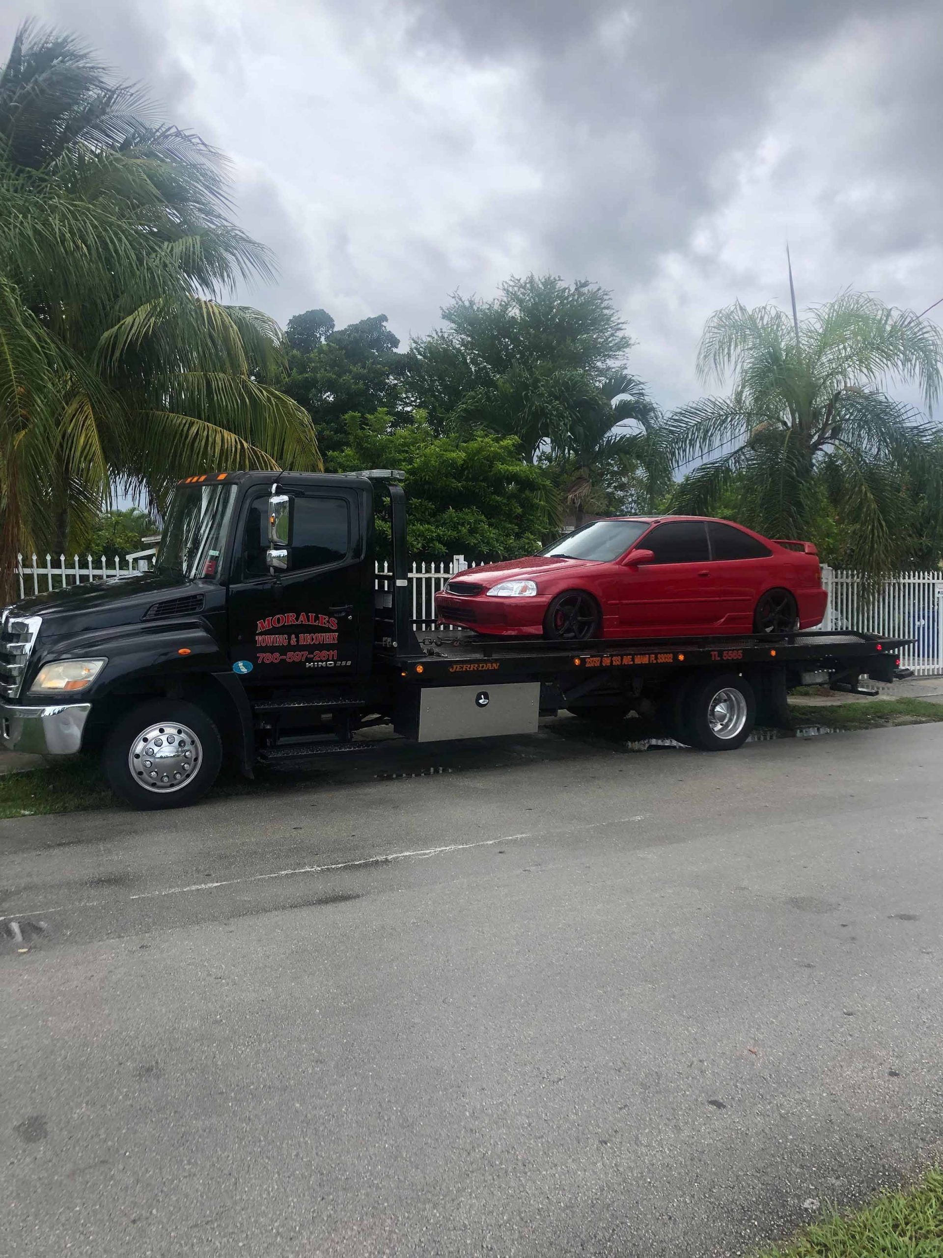 Black tow truck with red car on the flatbed, parked on a street. Overcast day.