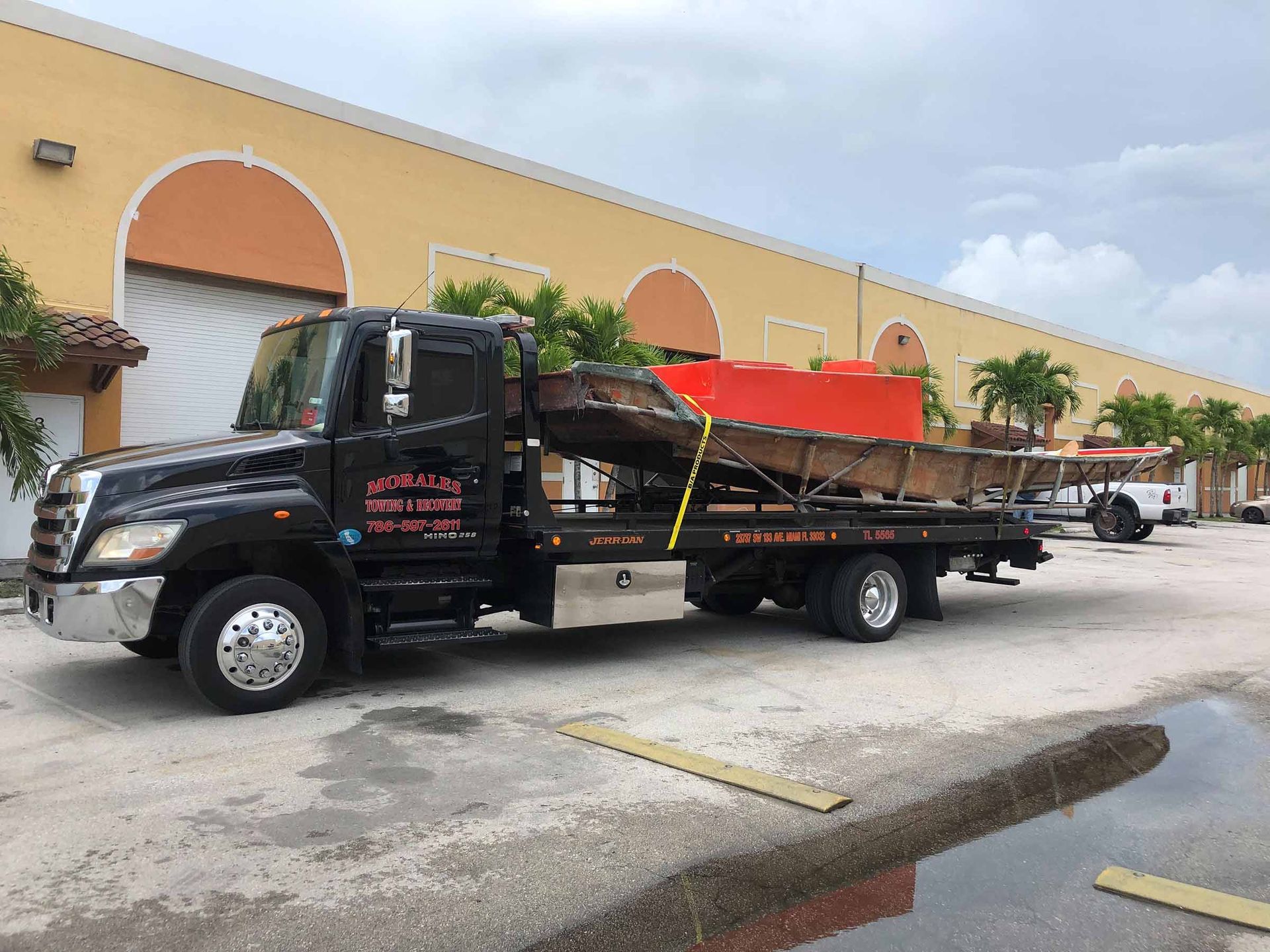 Black tow truck carrying a boat, parked in front of a building with arches.