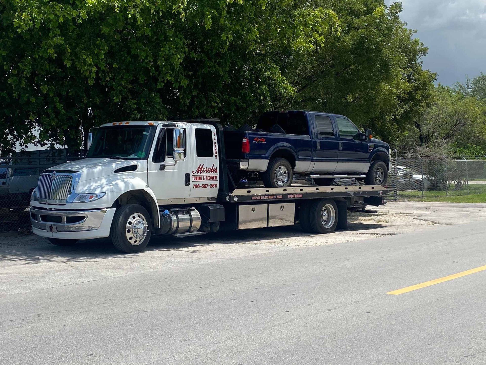 Tow truck carrying a blue pickup truck on a roadside, daytime.