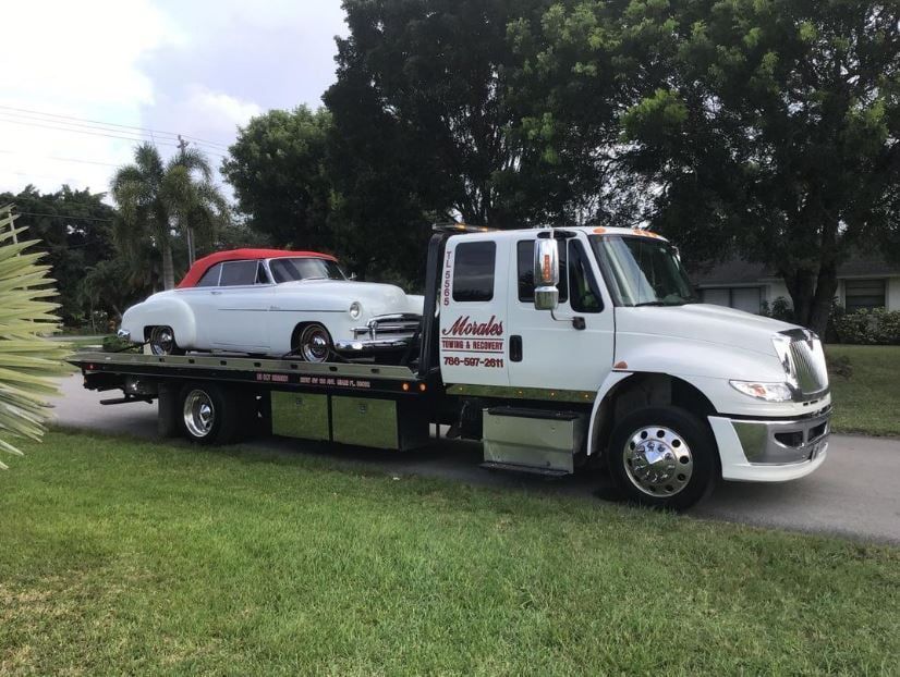 Tow truck hauling a white vintage car with a red convertible top on a residential street.