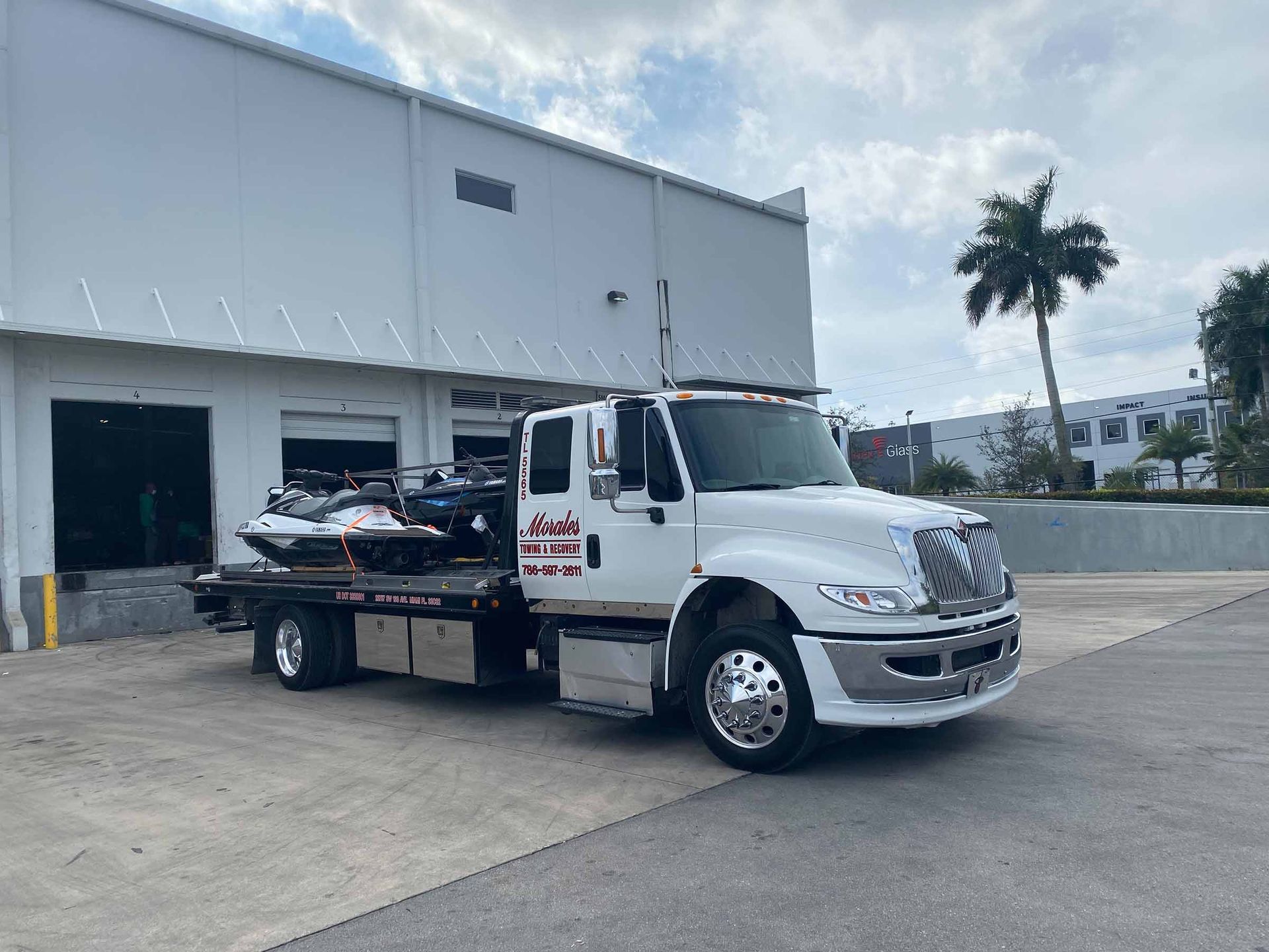 White tow truck with a personal watercraft on its flatbed, parked near a warehouse.