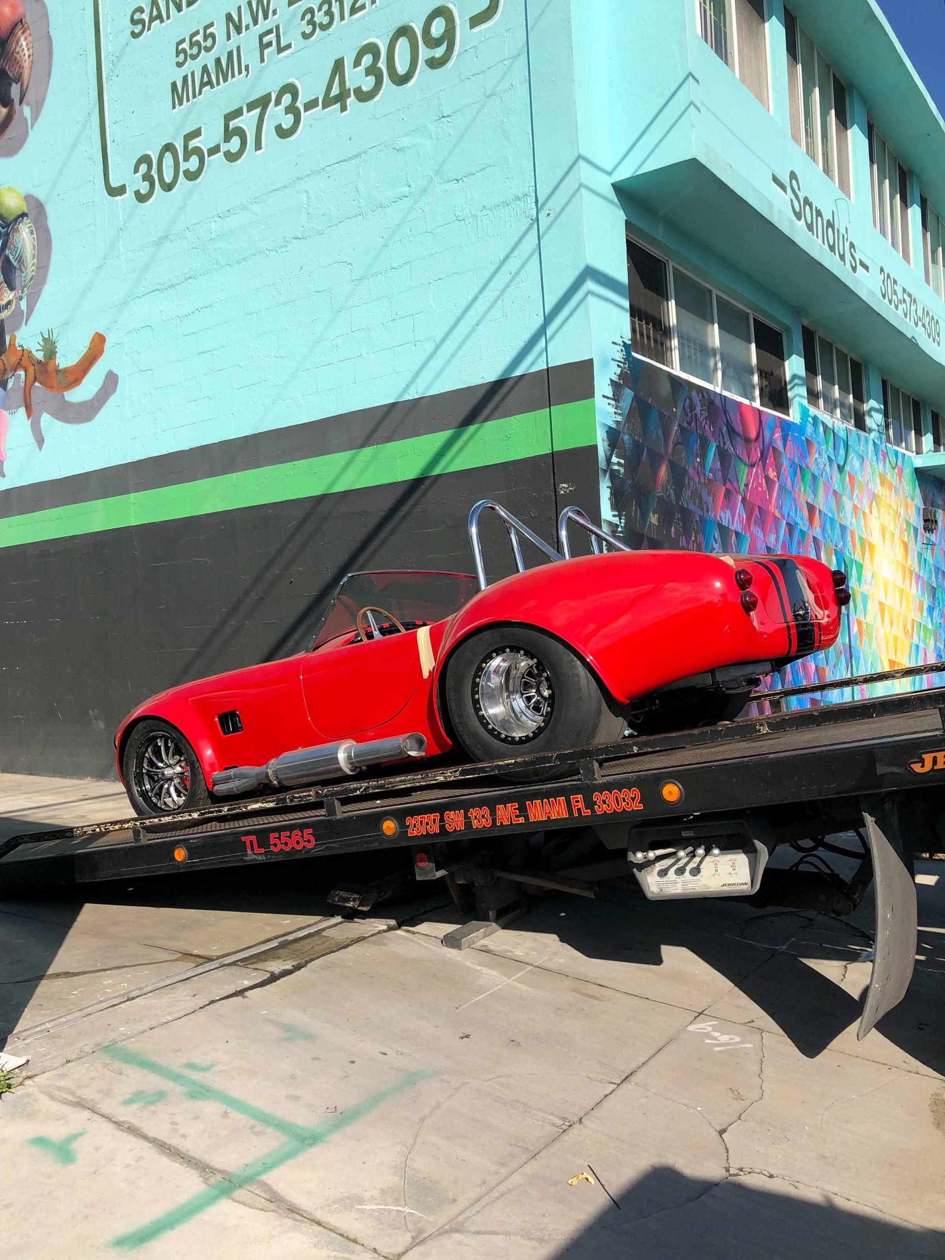 Red sports car on a tow truck, next to a teal building with graffiti in Miami.